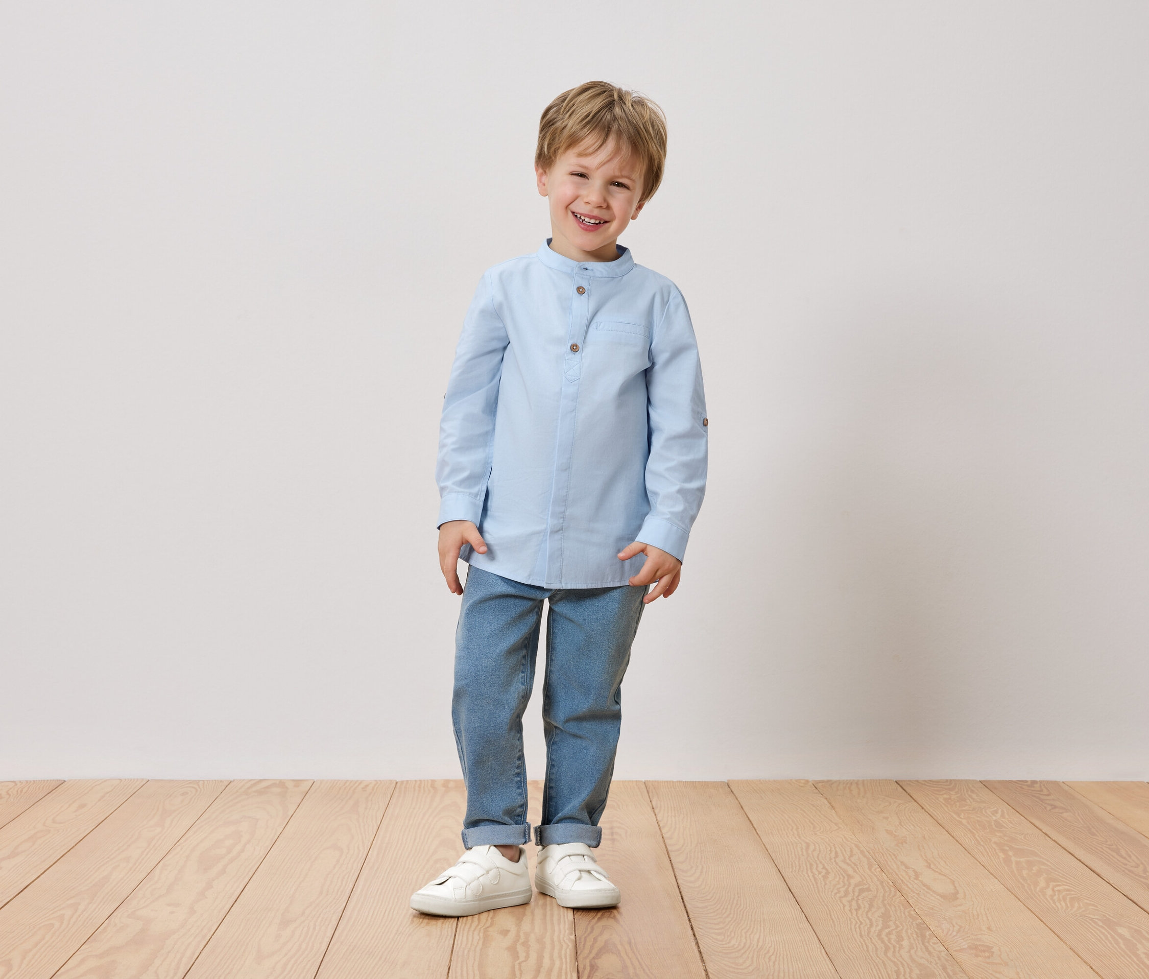 Ein lächelnder Junge mit blonden Haaren steht vor einer weißen Wand. Er trägt ein hellblaues Hemd, Jeans und weiße Schuhe.