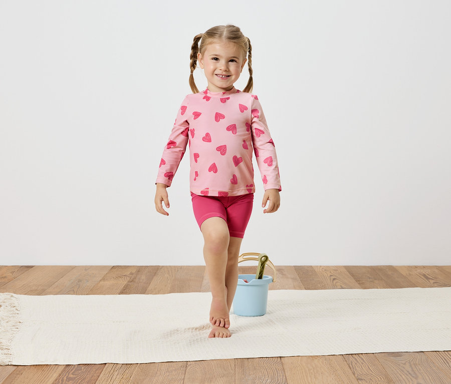 Mädchen mit Zöpfen, trägt ein rosa Herzchenoberteil und Shorts, balanciert auf einem weißen Teppich neben einem hellblauen Eimer.