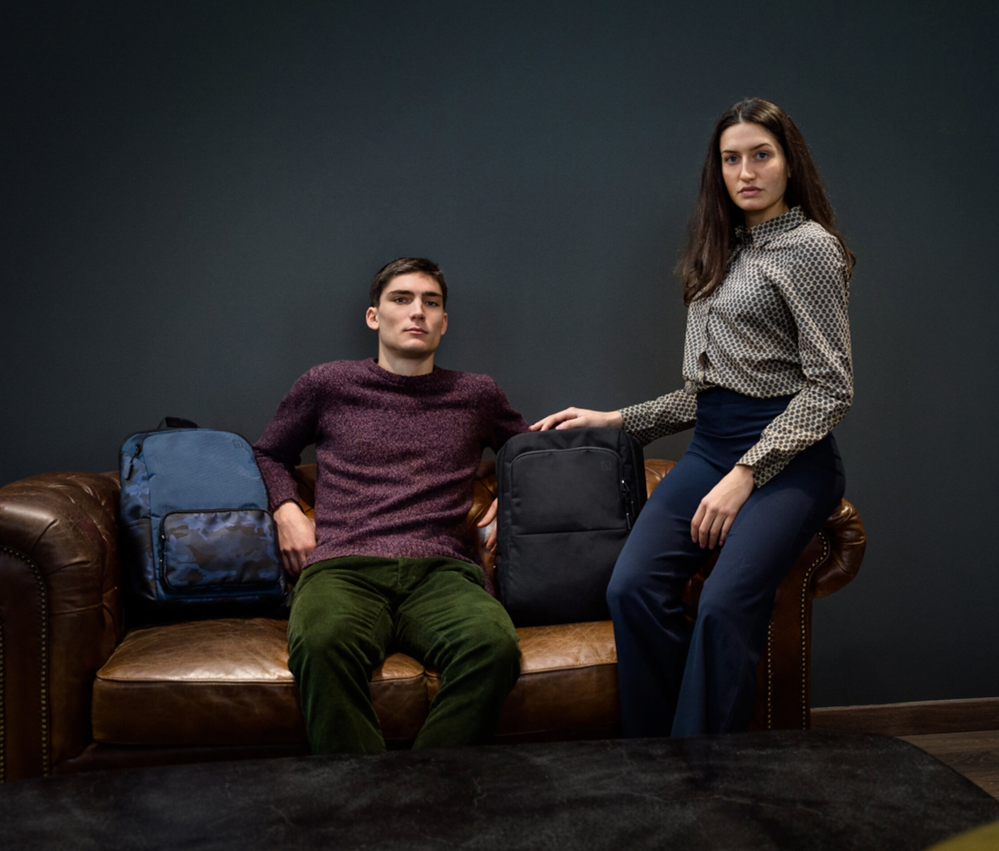 Zwei Personen sitzen auf einem Ledersofa, neben ihnen ein blauer und ein schwarzer Tucano PLANET Lunar Rucksack, 15,6".