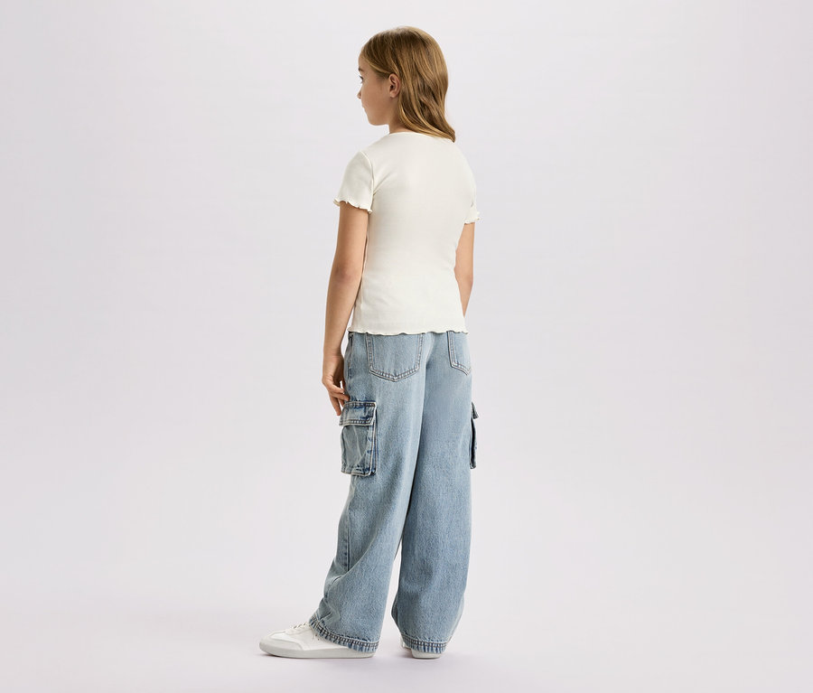 Mädchen von hinten, trägt ein kurzes, cremefarbenes Oberteil, weite Cargo-Jeans und weiße Turnschuhe vor einem weißen Hintergrund.