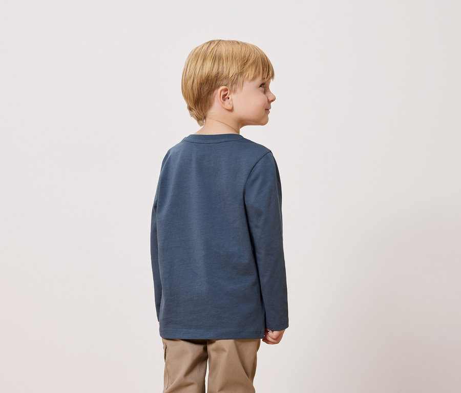 Ein blonder Junge steht mit dem Rücken zur Kamera und trägt ein blaues Langarmshirt und Khakihosen.