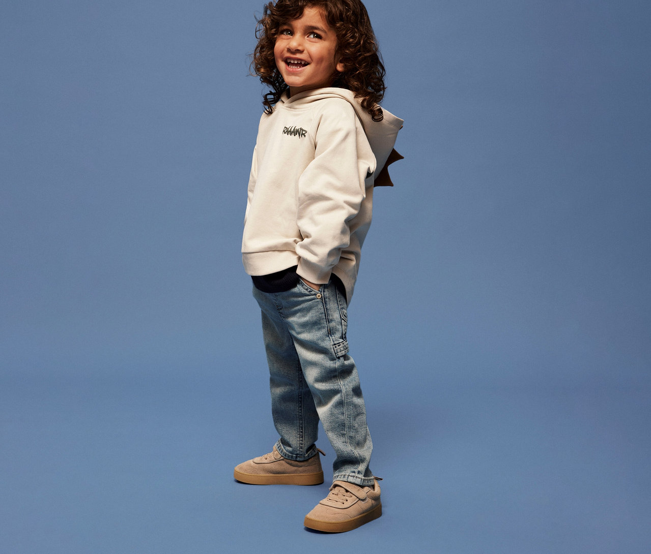 Junge mit lockigem Haar trägt ein NAME IT Kinder-Sweatshirt mit 3D-Dino-Applikation, Jeans und Turnschuhe vor blauem Hintergrund.