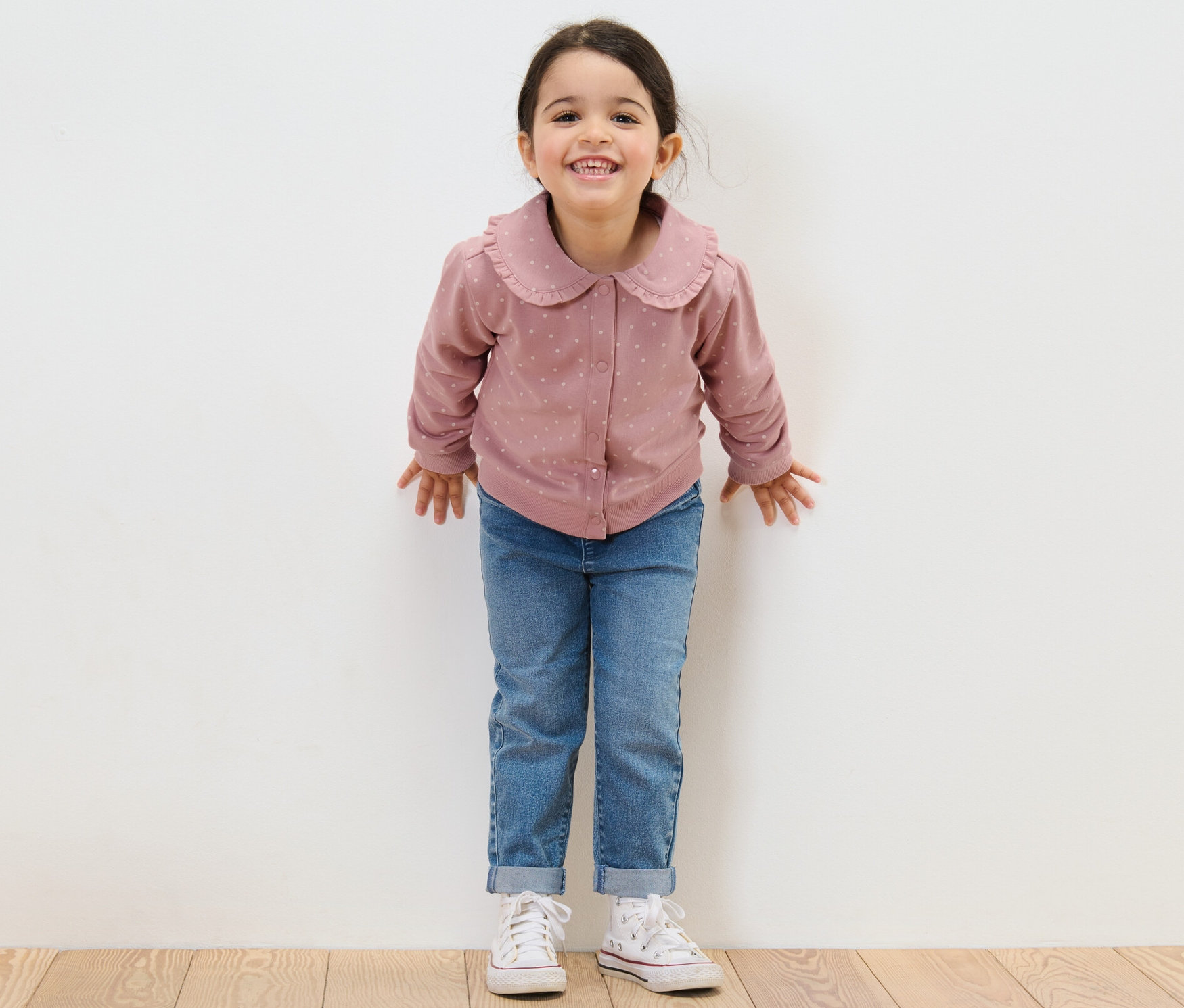 Lächelndes Mädchen steht vor einer weißen Wand und trägt Jeans und eine Kinder-Sweatjacke mit Volant.