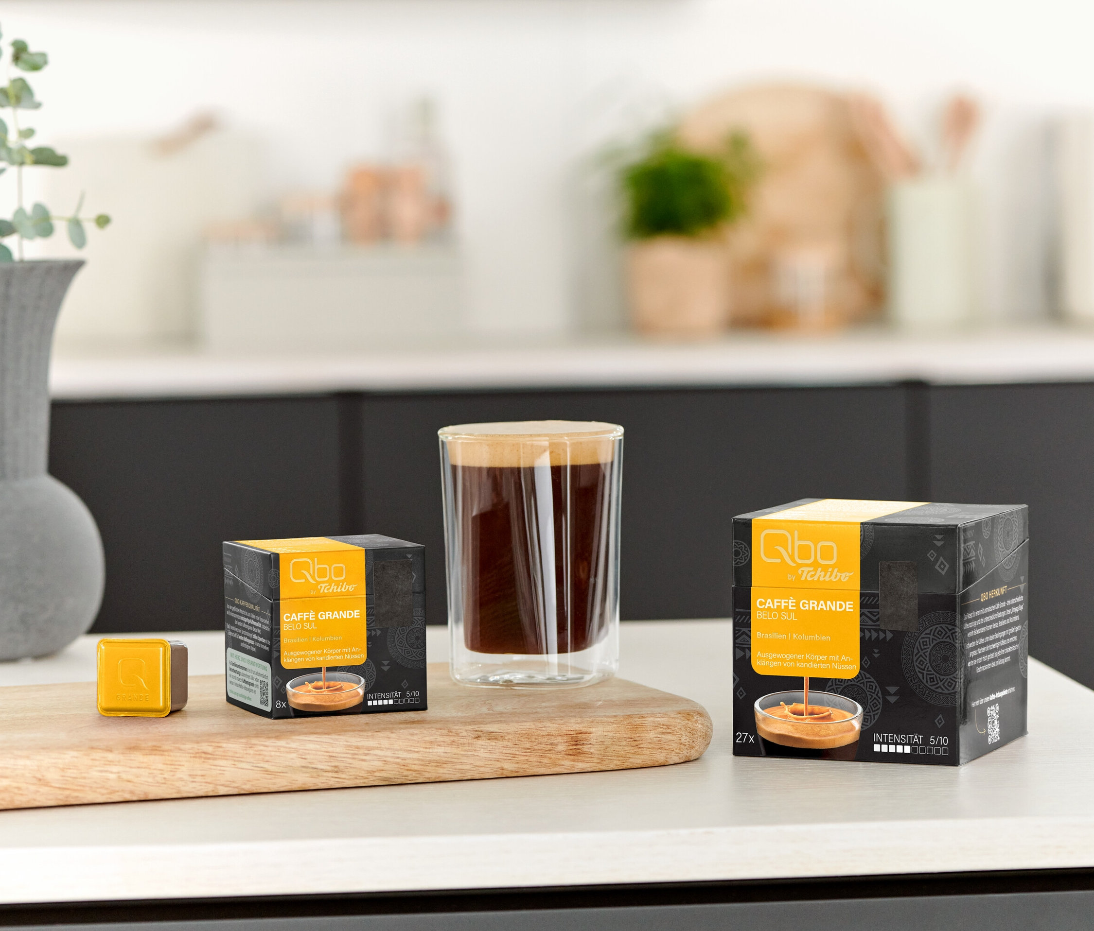 Ein Glas Kaffee steht neben einer Qbo Caffè Grande BELO SUL - 8 Kapsel Packung und einer einzelnen Kapsel auf einem Holzbrett.