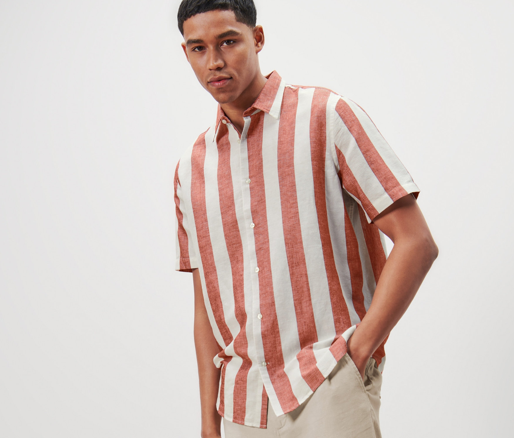 Mann posiert in einem gestreiften Kurzarmhemd mit Leinenanteil und hellen Hosen.
