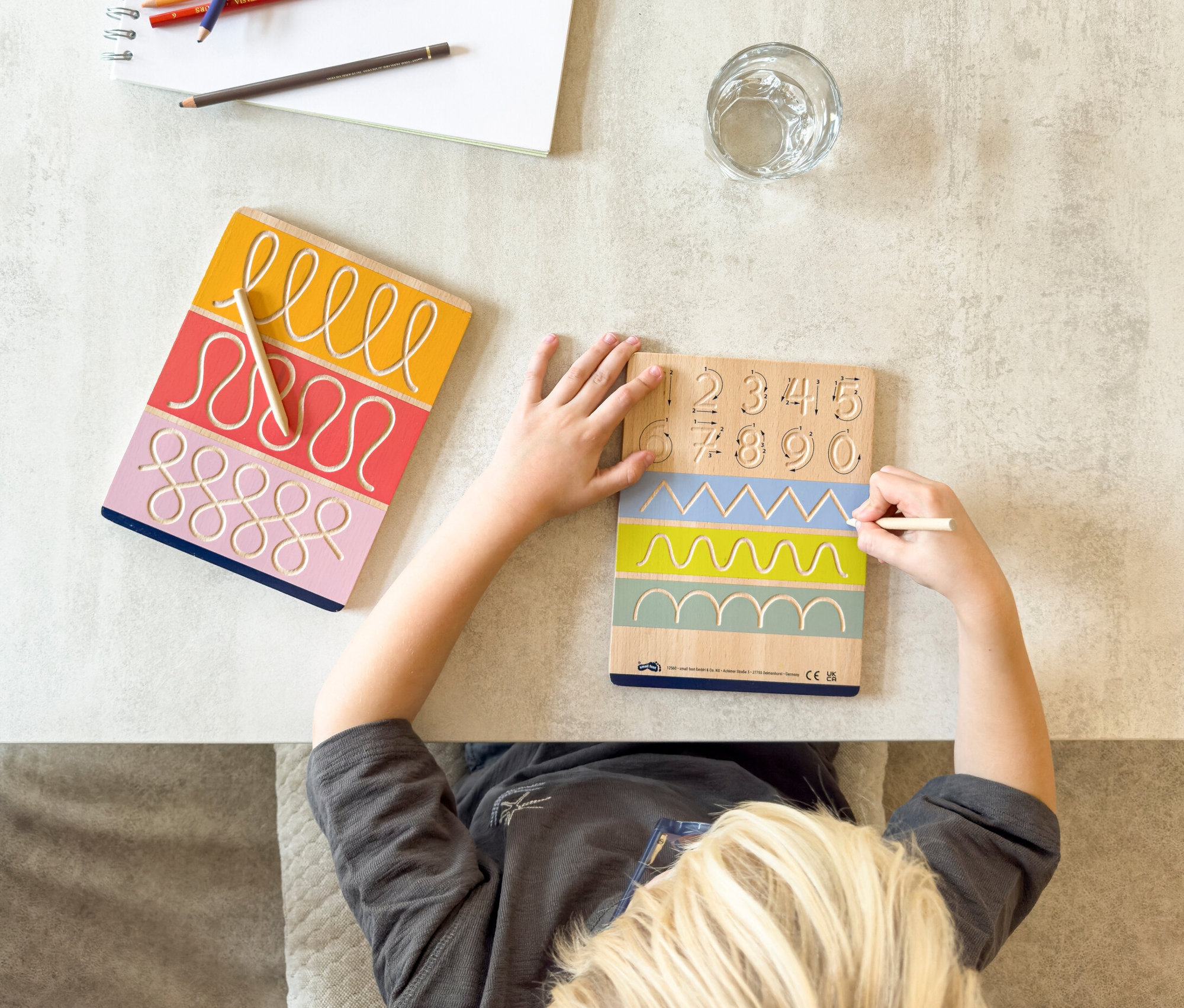 Junge schreibt auf eine Holztafel mit Zahlen und Formen, daneben liegt eine small foot Schreibtafel »Educate« und ein Skizzenblock.