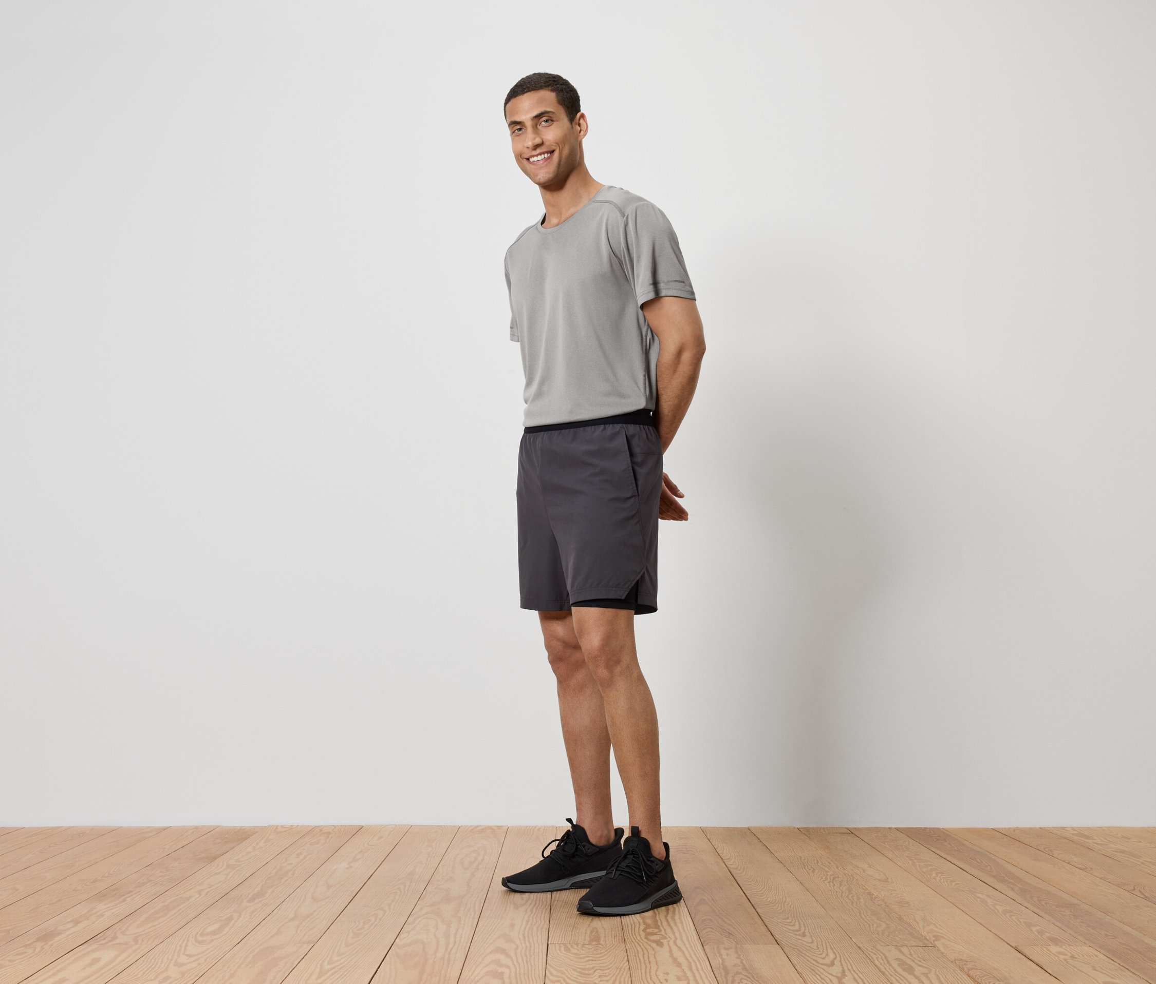 Mann mit grauem T-Shirt, Shorts und Turnschuhen steht auf einem Holzboden vor einer weißen Wand.