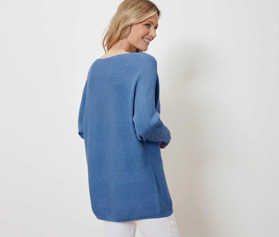 Blonde Frau lächelt über die Schulter und trägt einen blauen Feinstrickpullover und weiße Hosen.