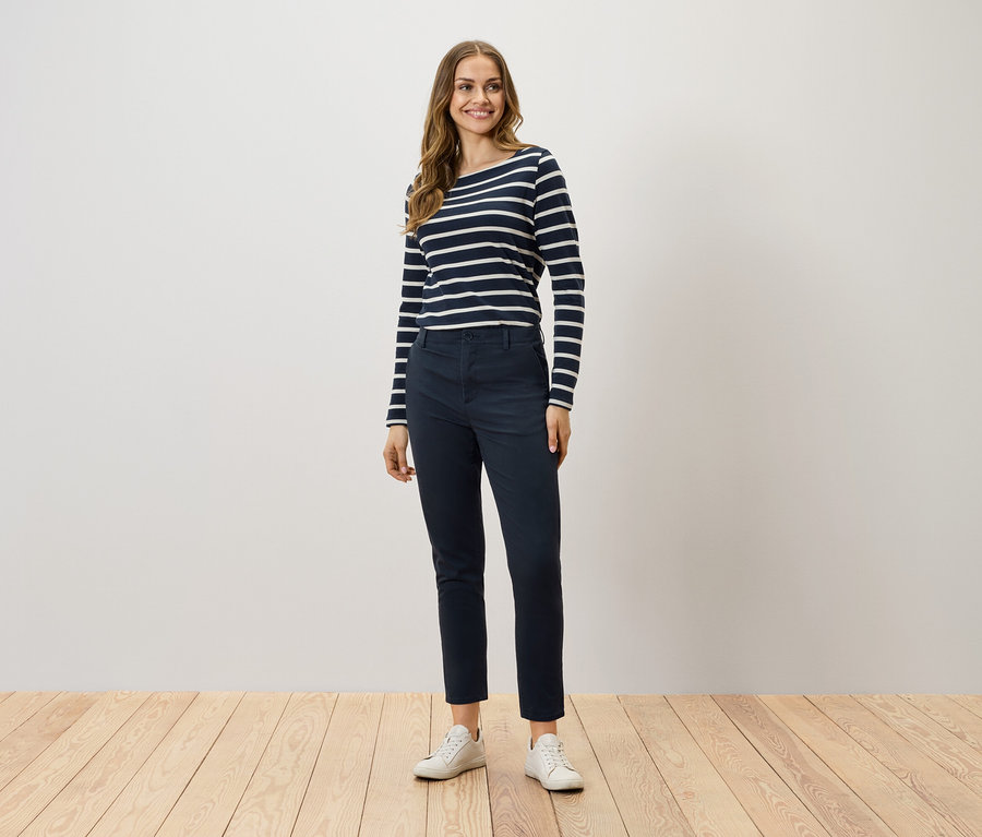 Eine Frau steht und trägt ein langärmliges Streifenshirt, dunkelblaue Hosen und weiße Turnschuhe.