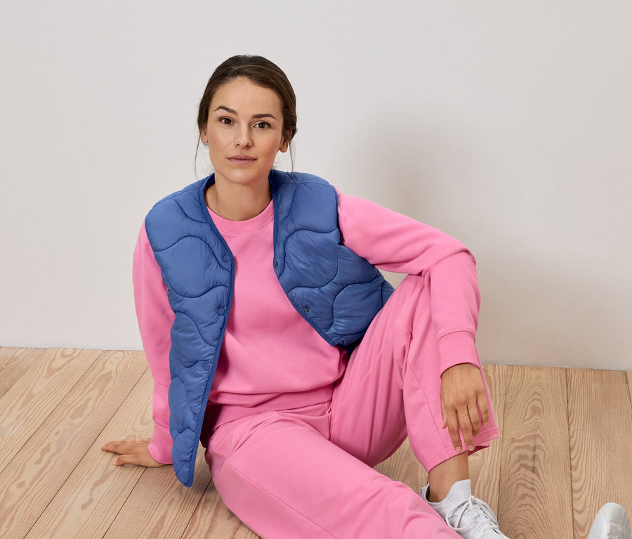 Frau sitzt in rosa Trainingsanzug und blauer Weste auf dem Boden.