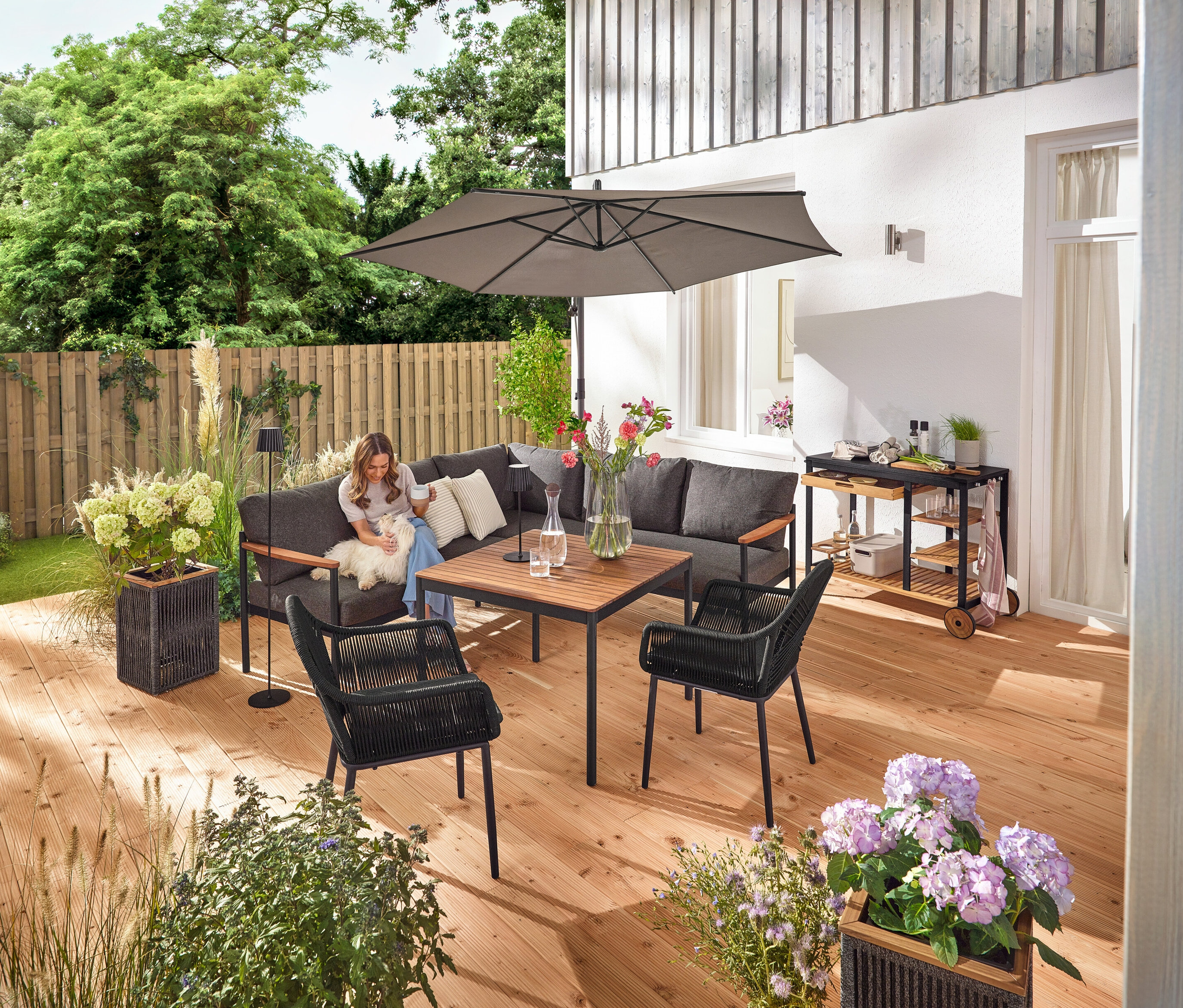 Auf einer Terrasse sitzt eine Frau mit einem Hund auf einer Dining-Lounge-Ecke »Elin«. Ein Grillwagen mit Polyzement-Arbeitsfläche, ein Pflanztopf mit Textilgeflecht, eine LED-Akku-Outdoor-Stehleuchte, eine LED-Akku-Outdoor-Tischleuchte, zwei Outdoor-Kissen mit Leinen-Haptik und ein Gartenstuhl mit Textil-Kreuzgeflecht sind ebenfalls zu sehen.