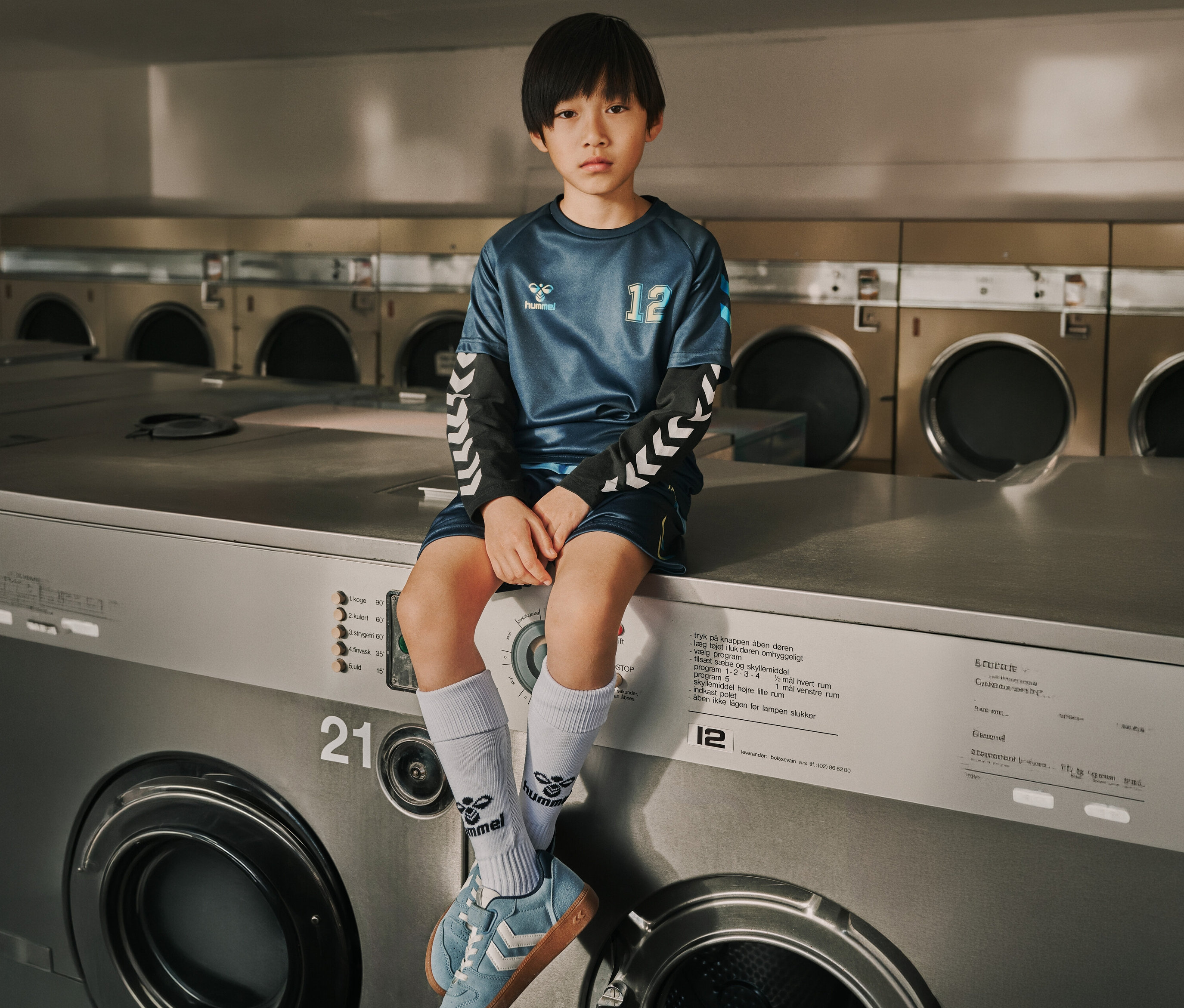 Junge im marineblauen hummel® Kinder-Trikot-Set HMLSHIMMER sitzt auf einer Waschmaschine.