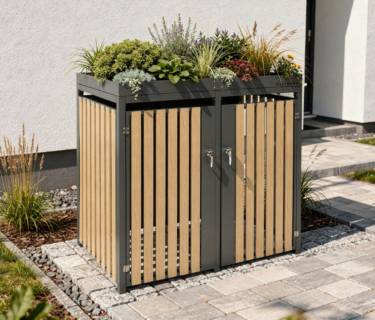 Eine Mülltonnenbox mit Blumenkasten, 2er, steht auf einem gepflasterten Weg vor einem Haus. Auf der Oberseite befinden sich verschiedene Pflanzen.