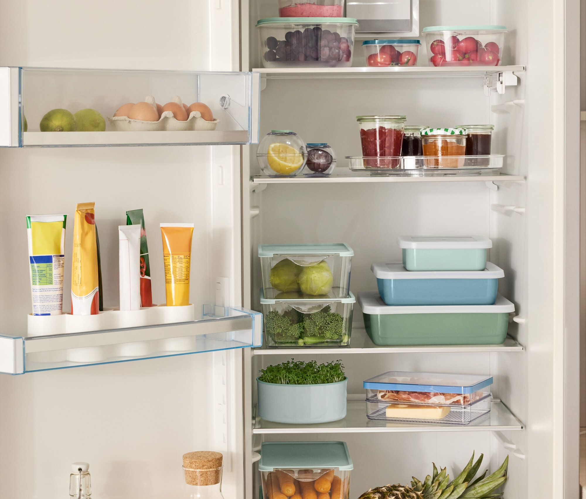 Offener Kühlschrank gefüllt mit verschiedenen Lebensmitteln, darunter 4 Aufbewahrungsboxen, 2 Frischhaltekugeln, 3 Emaille-Auflaufformen mit Deckel und einem Drehteller.