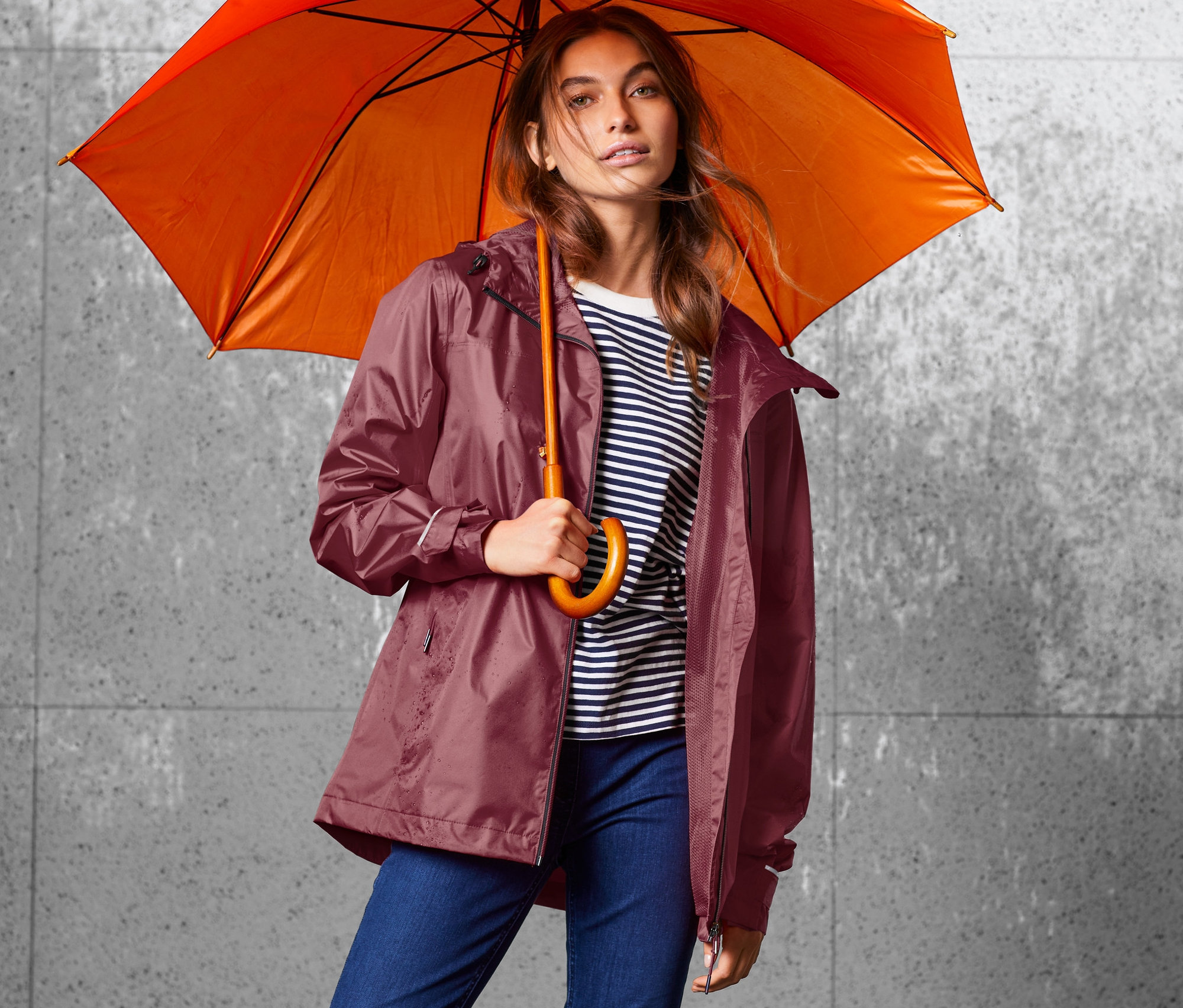 Frau hält orangefarbenen Regenschirm und trägt weinrote Regenjacke.