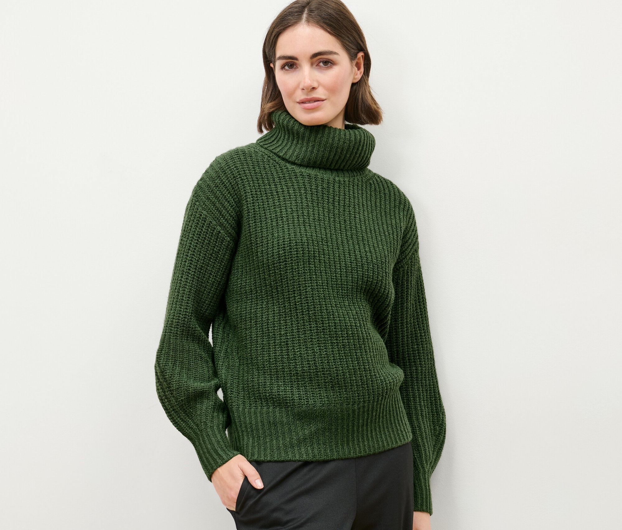 Frau posiert vor hellem Hintergrund in einem Grobstrickpullover mit Rollkragen.