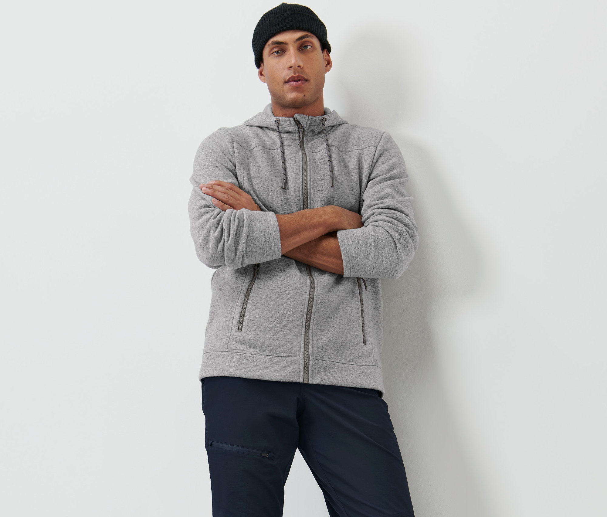 Mann posiert mit verschränkten Armen in einer grauen Strickfleece-Jacke und einer schwarzen Mütze.