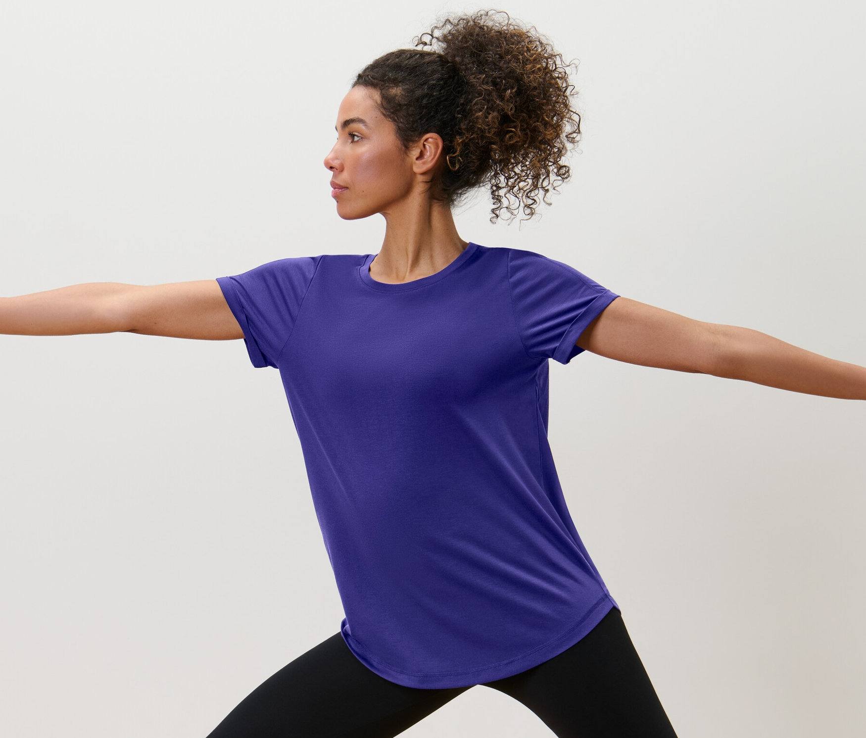 Frau in schwarzen Leggings und lila Longshirt steht mit ausgestreckten Armen.