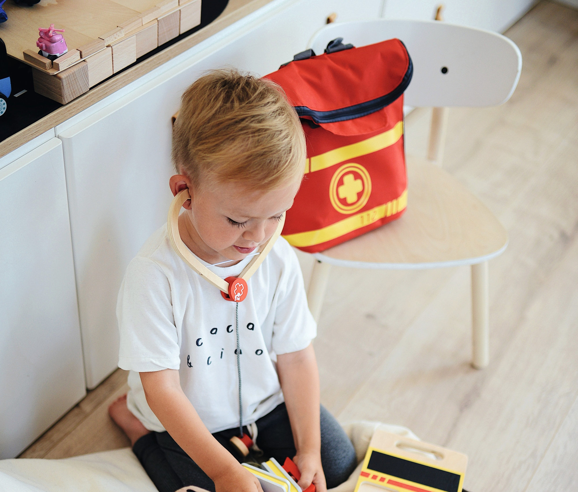 Ein Junge spielt mit einem Teddybär und einem Arztkoffer, trägt ein Stethoskop und daneben liegt ein small foot Notarzt-Rucksack.