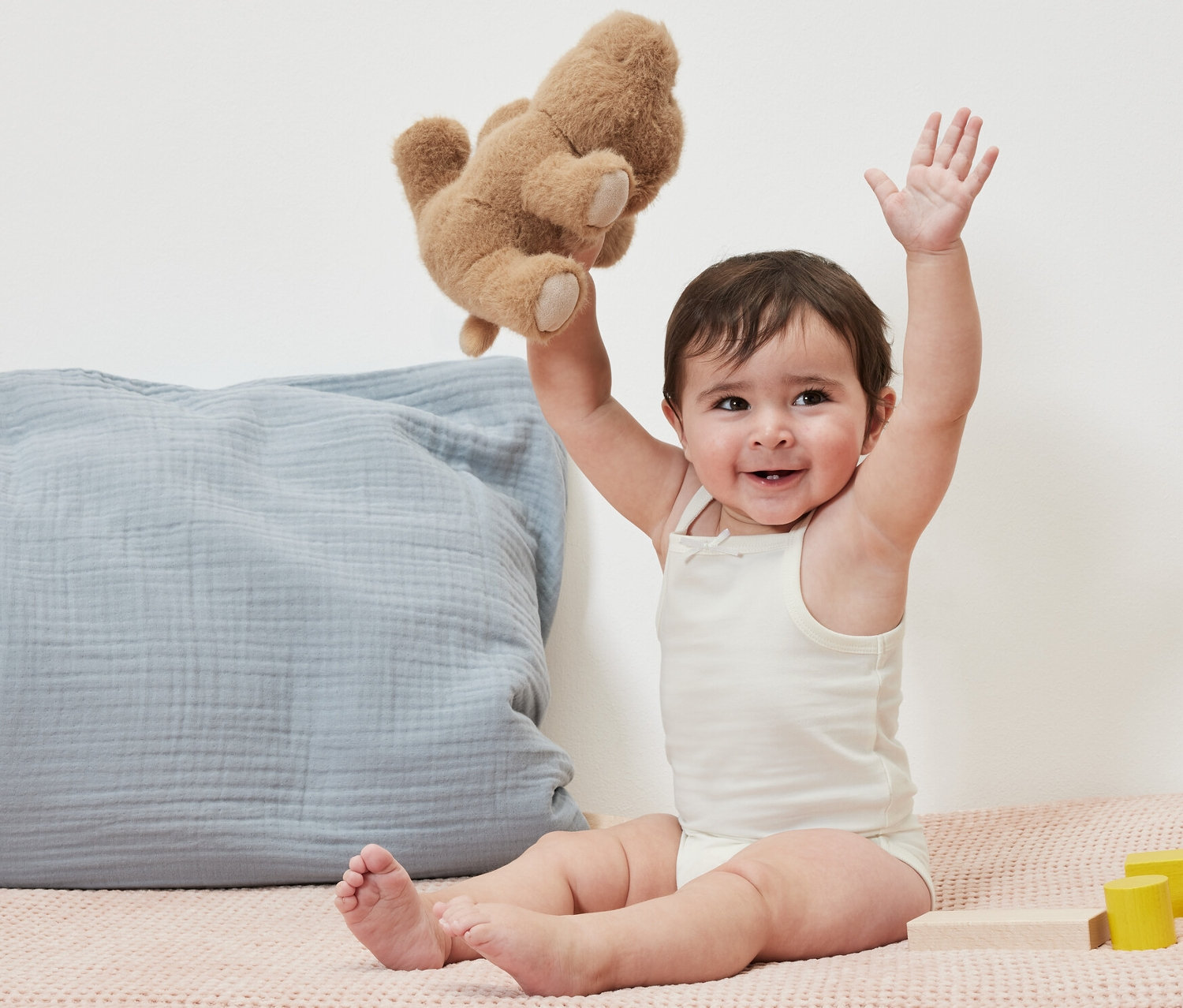 Ein lächelndes Baby sitzt und hält einen Teddybär hoch. Es trägt 5 Baby-Bodys.