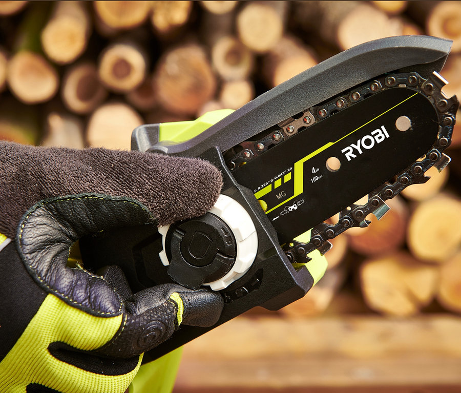 Eine Hand in einem Handschuh hält eine bürstenlose Akku-Einhand-Kettensäge Ryobi 18 V One+ vor einem Holzstapel.