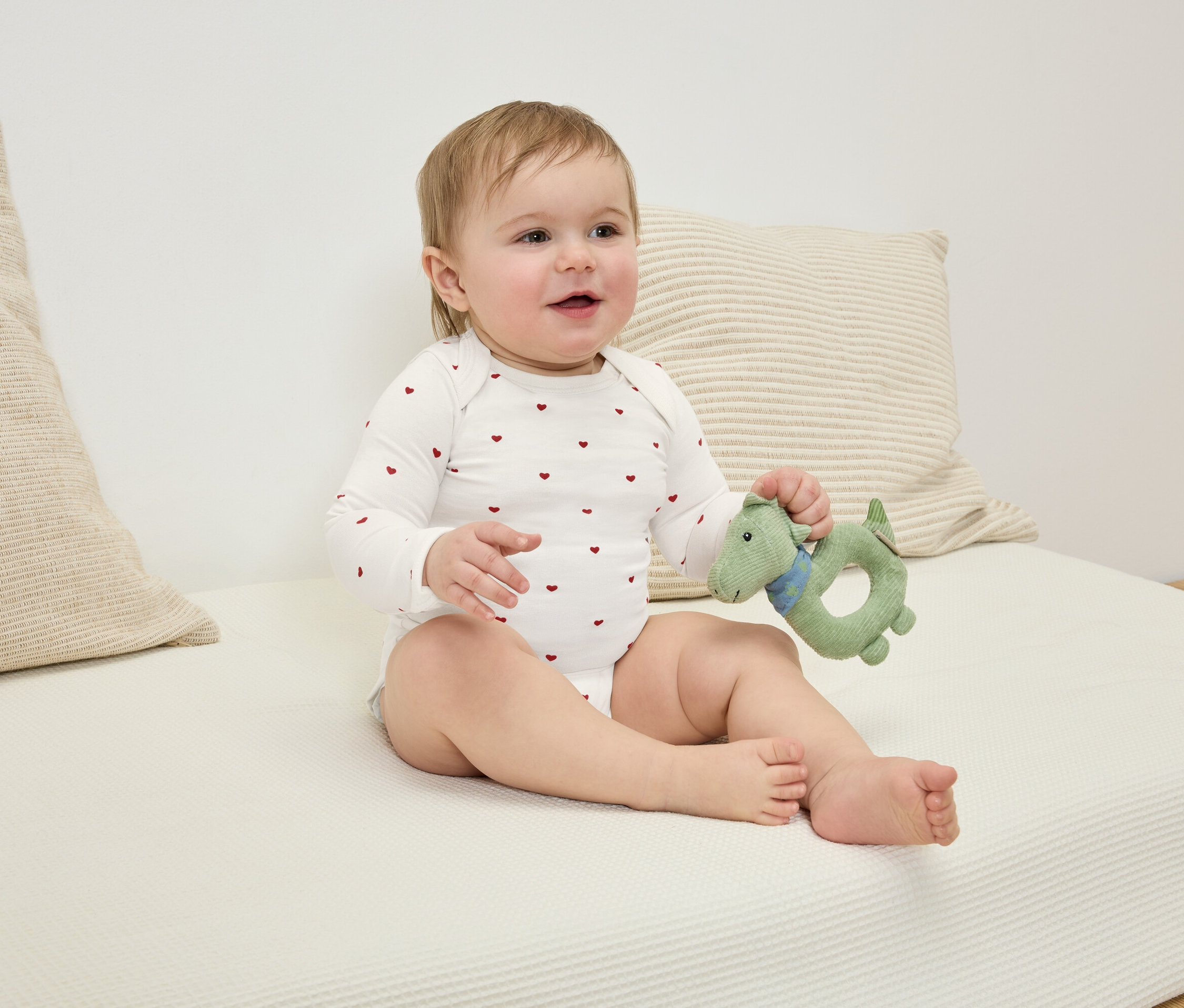 Sitzendes Baby in einem Body mit roten Herzen hält ein grünes Dinosaurier-Spielzeug.