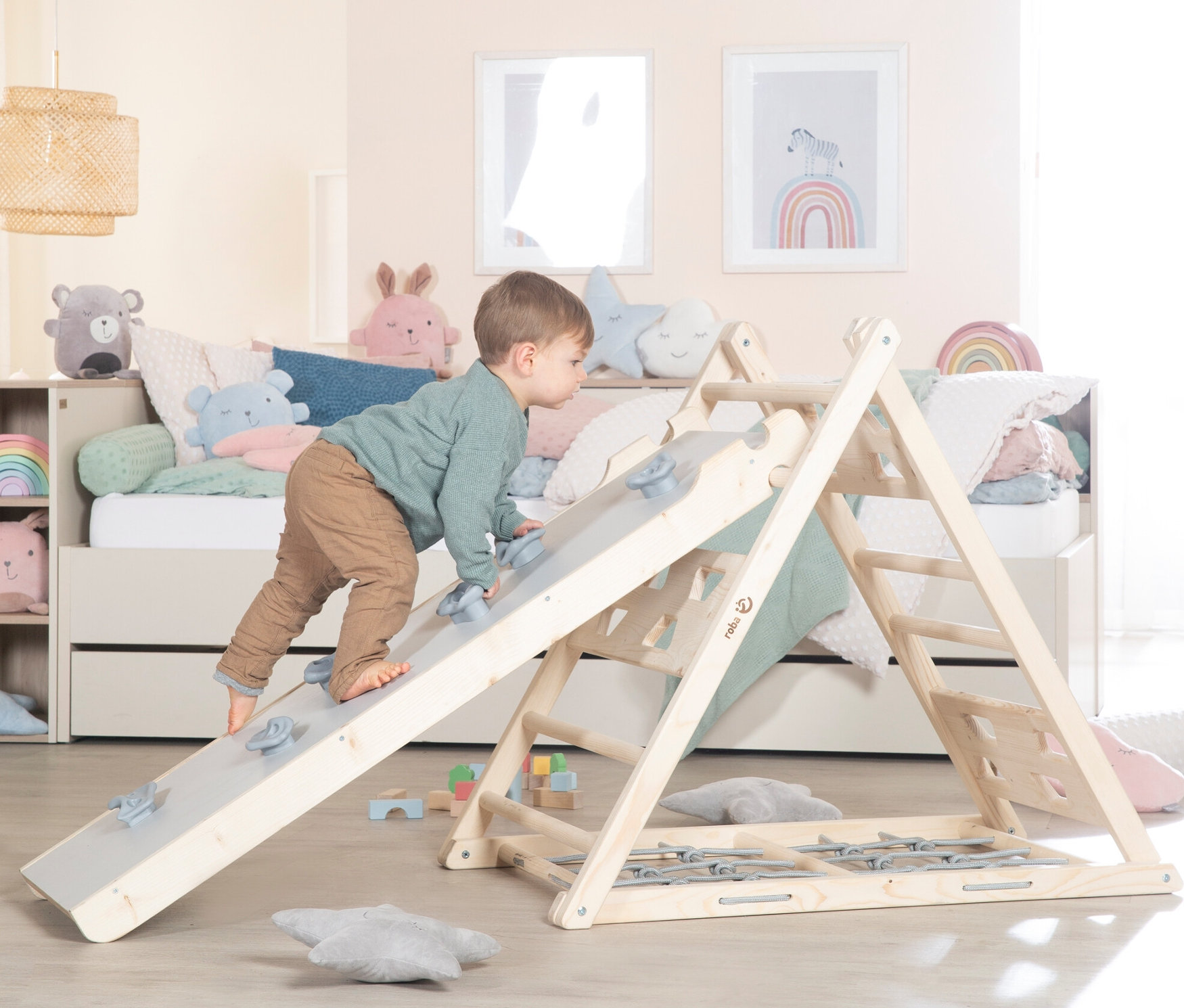 Junge klettert auf roba Rutschbrett Klettergerüst mit grauer Rutsche. Im Hintergrund sind ein Bett mit Stofftieren und Bilder an der Wand zu sehen.