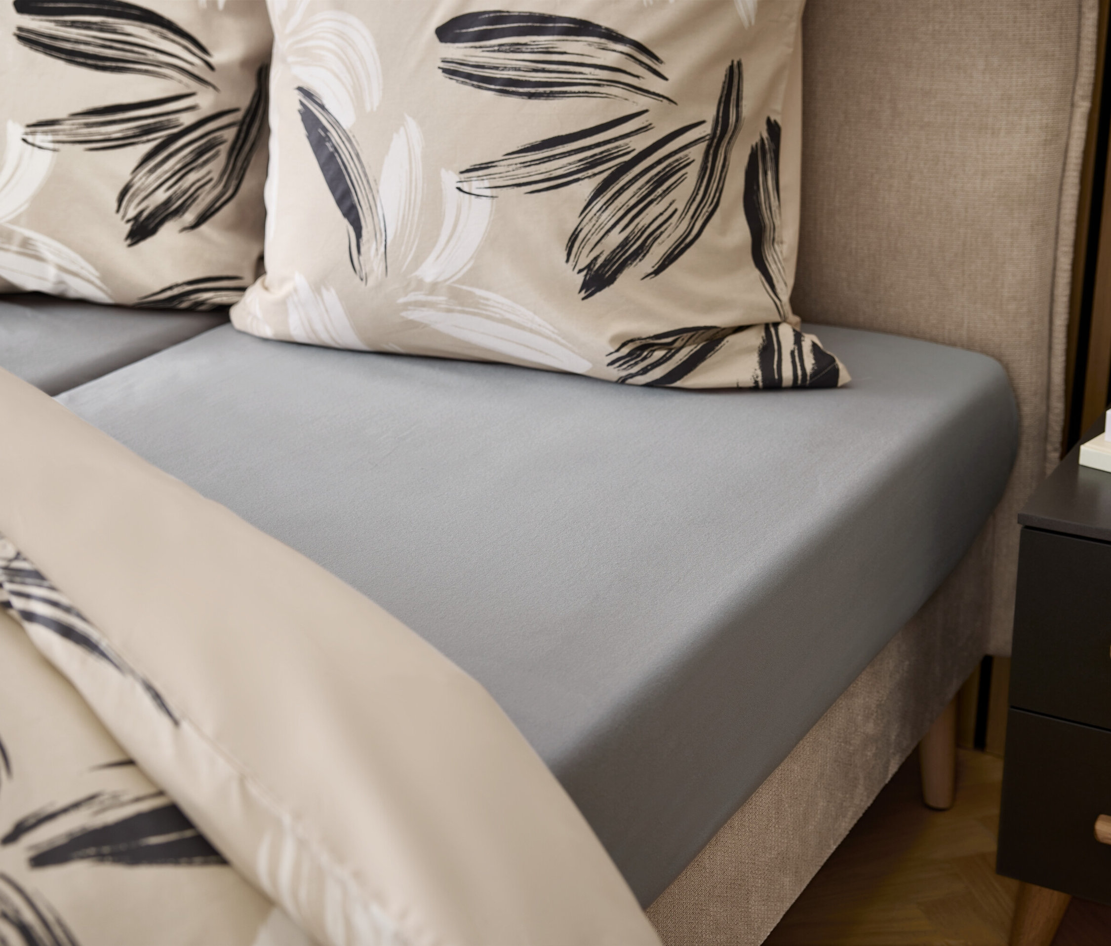 Detailansicht eines grauen Bettes mit floralen Kissen und Nachttisch.