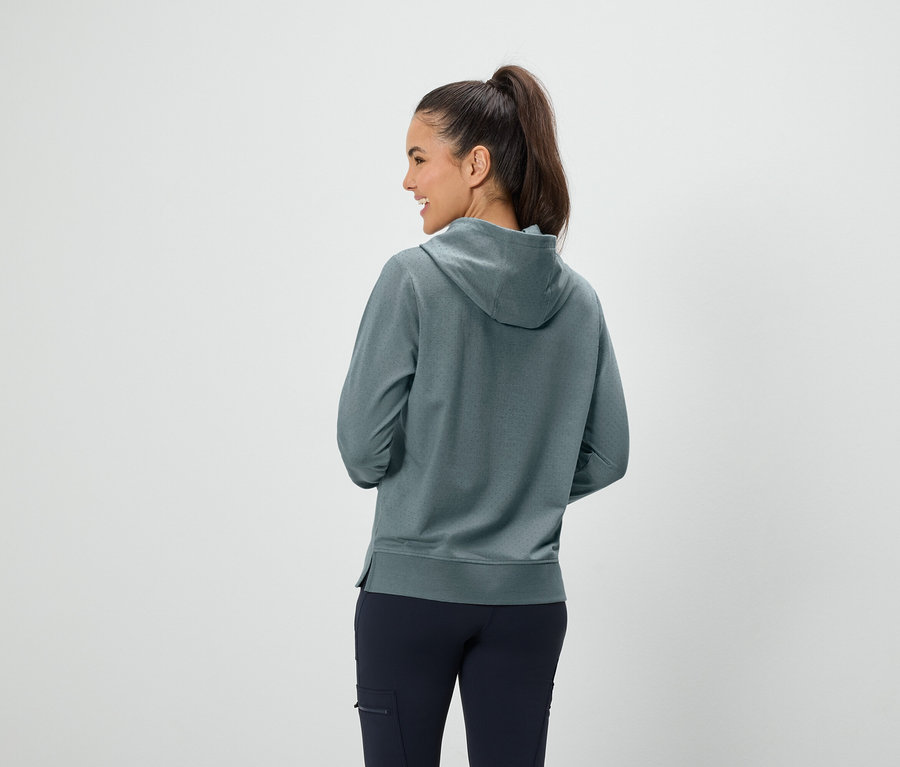 Eine Frau mit braunen Haaren im Pferdeschwanz steht mit dem Rücken zur Kamera und trägt einen blaugrünen Kapuzenpullover und dunkelblaue Hosen.