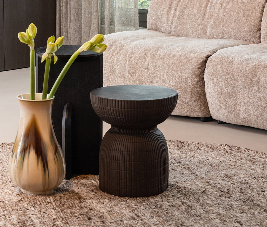 Eine Vase mit Blumen steht auf dem Woood Teppich »Efia« neben einem braunen Hocker und einem beigen Sofa.
