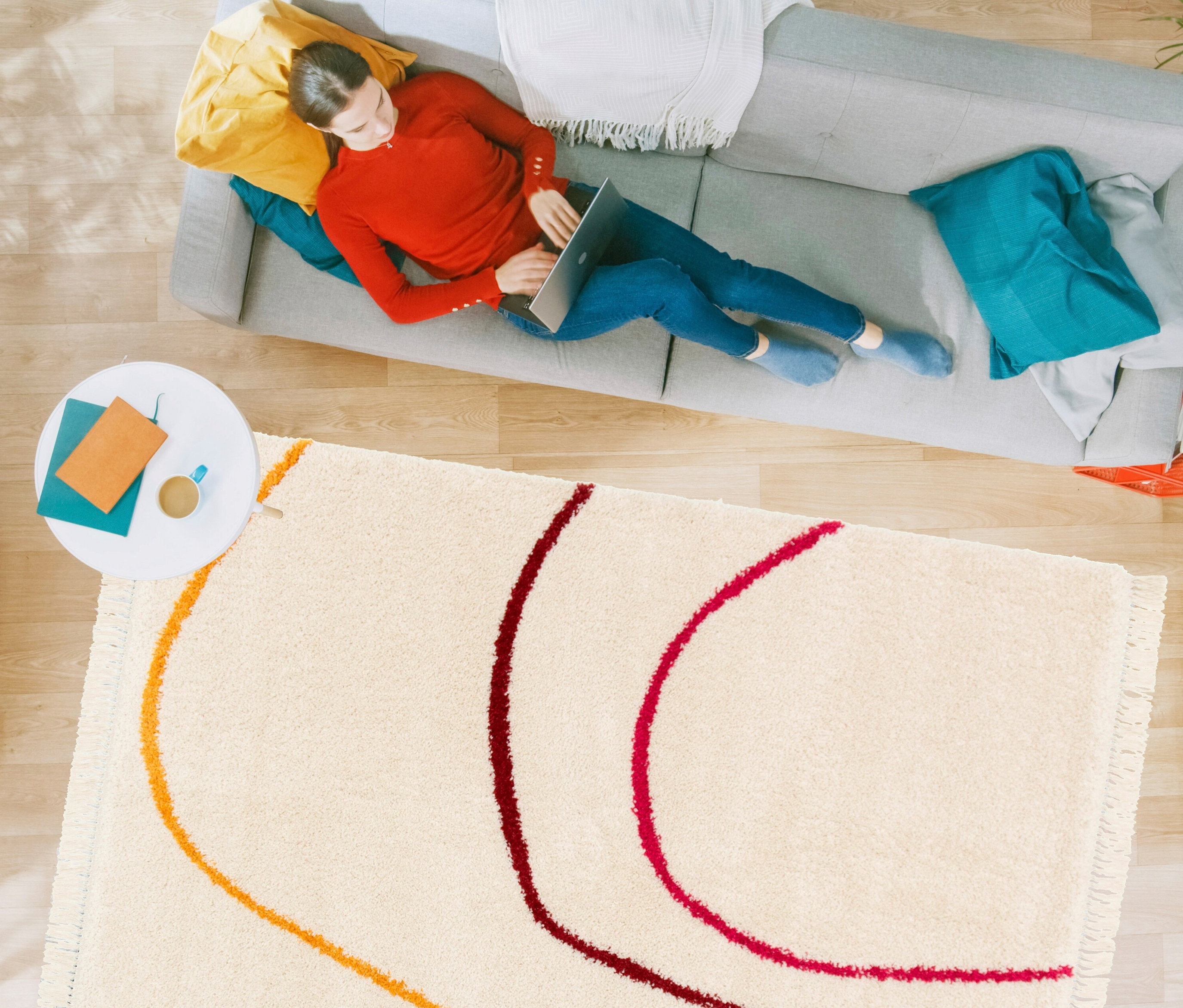 Frau liegt auf grauem Sofa und benutzt Laptop. Im Vordergrund liegt ein beiger Teppich »Mirage« mit bunten, wellenförmigen Linien.