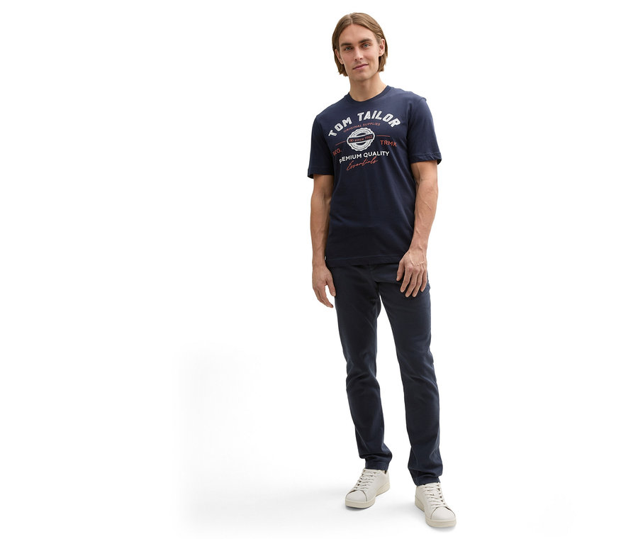 Mann in Hose und Tom Tailor T-Shirt mit Logo, Sky Captain Blue und weißen Schuhen steht vor einem weißen Hintergrund.