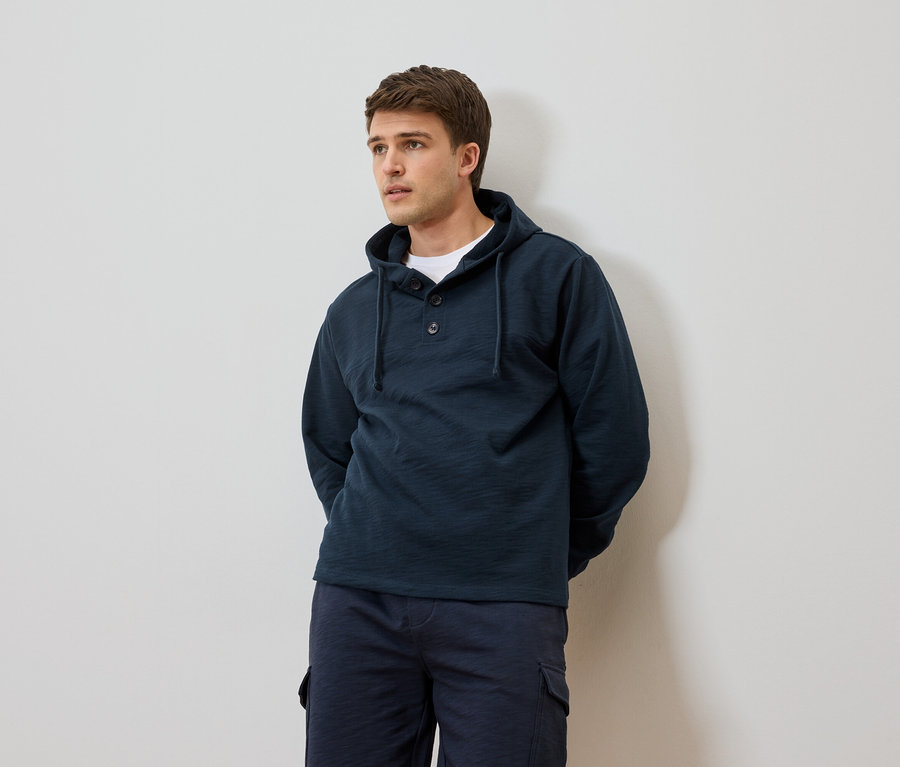 Mann mit dunkelblauem Kapuzenpullover und Shorts vor einer weißen Wand.