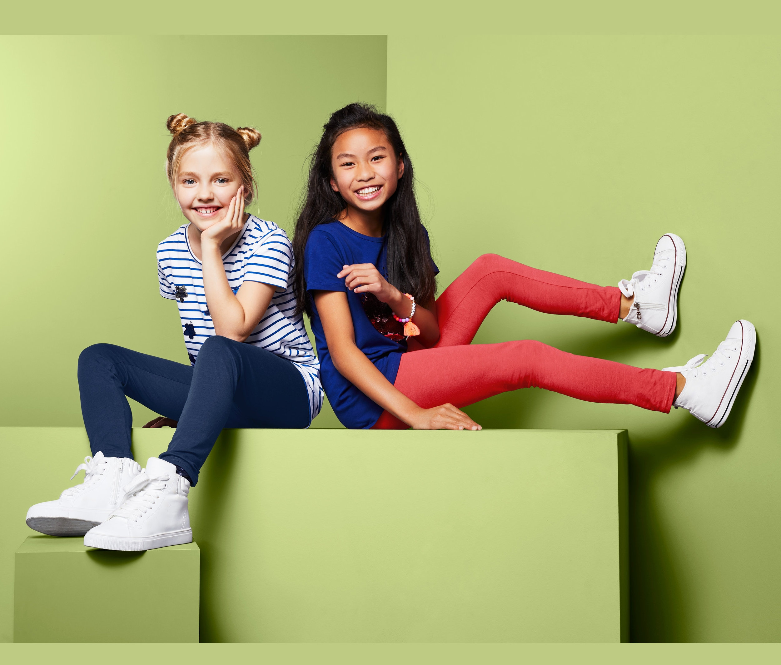 Zwei Mädchen sitzen auf einem grünen Block, eines in einem gestreiften Shirt und dunkelblauen Leggings, das andere in einem blauen Shirt und roten Leggings, beide tragen weiße Schuhe. 2 Leggings.