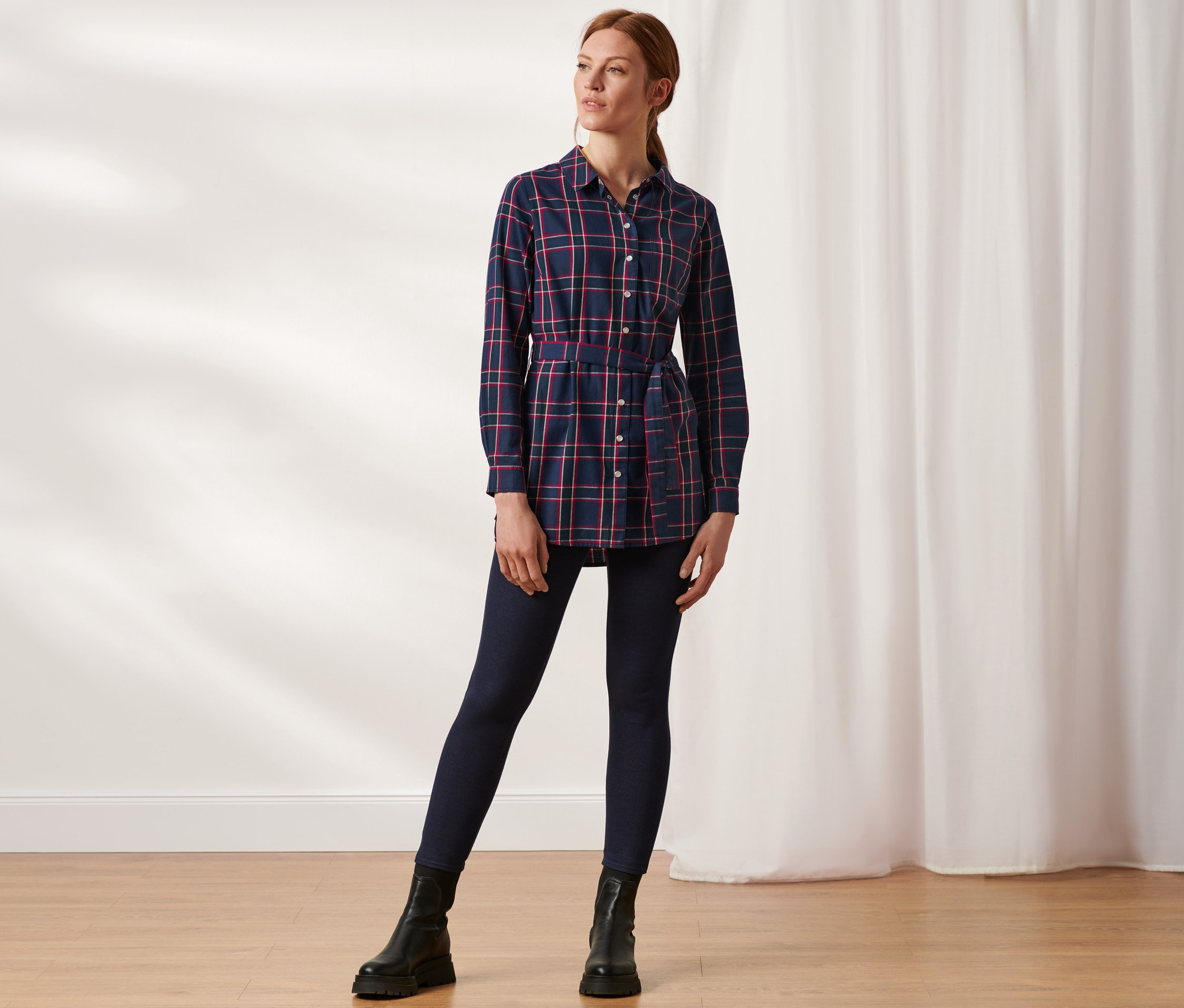 Frau posiert in karierter Longbluse aus Baumwoll-Flanell, dunkelblauen Jeans und schwarzen Stiefeln.