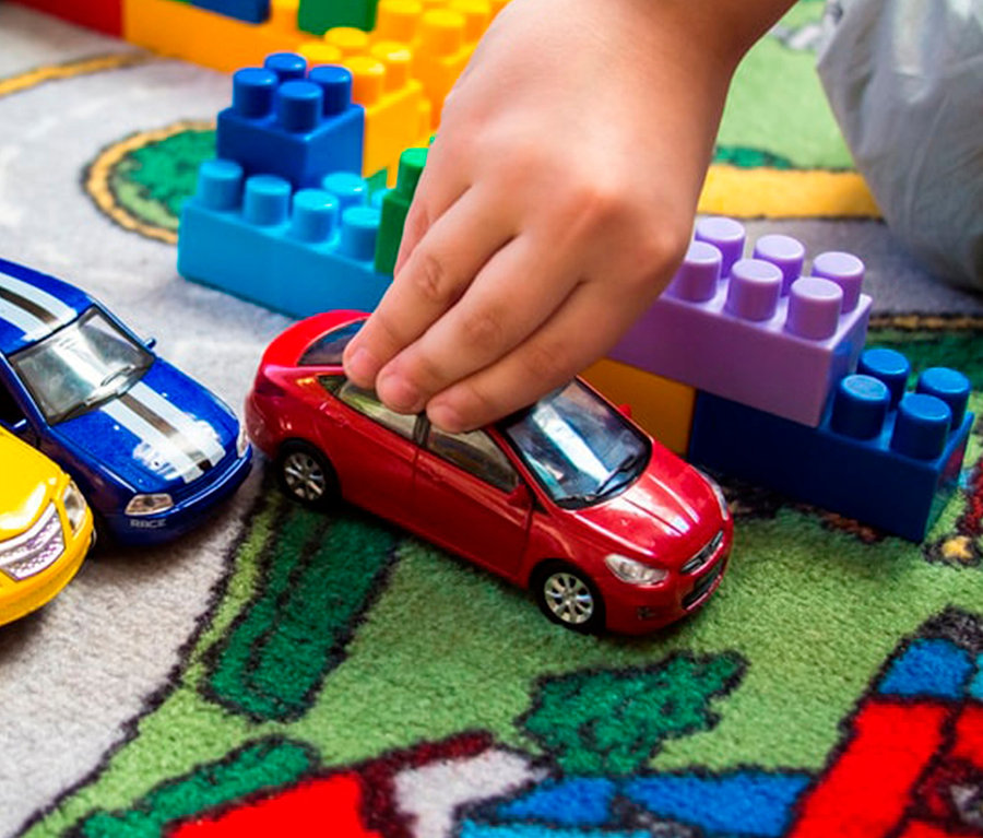 Kinderhand spielt mit rotem Spielzeugauto auf Spielteppich STREETS. Bunte Bauklötze und weitere Autos liegen umher.