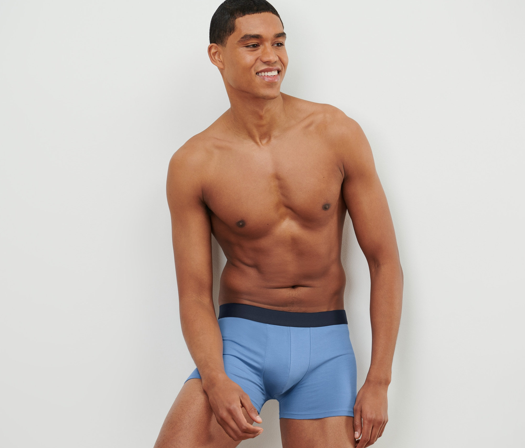 Ein Mann lächelt und lehnt an einer Wand. Er trägt blaue Slipboxer.