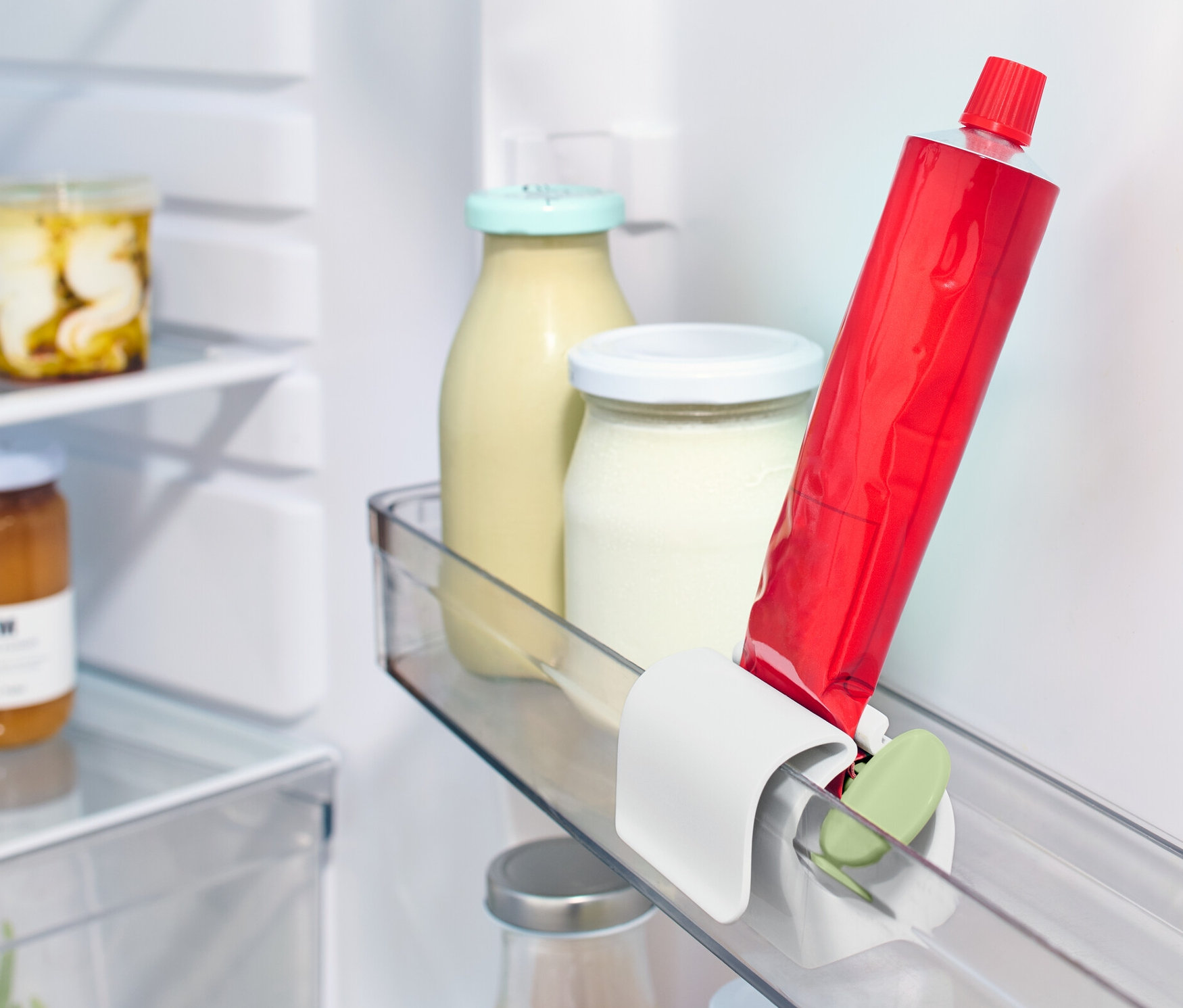 Eine rote Tube steckt in einem Tubenpresser mit Kühlschrankclip im Kühlschrankregal neben anderen Lebensmitteln.