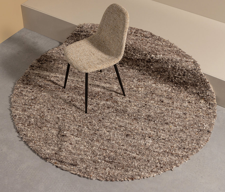 Ein beiger Stuhl steht auf dem braunen, runden Woood Teppich Efia.