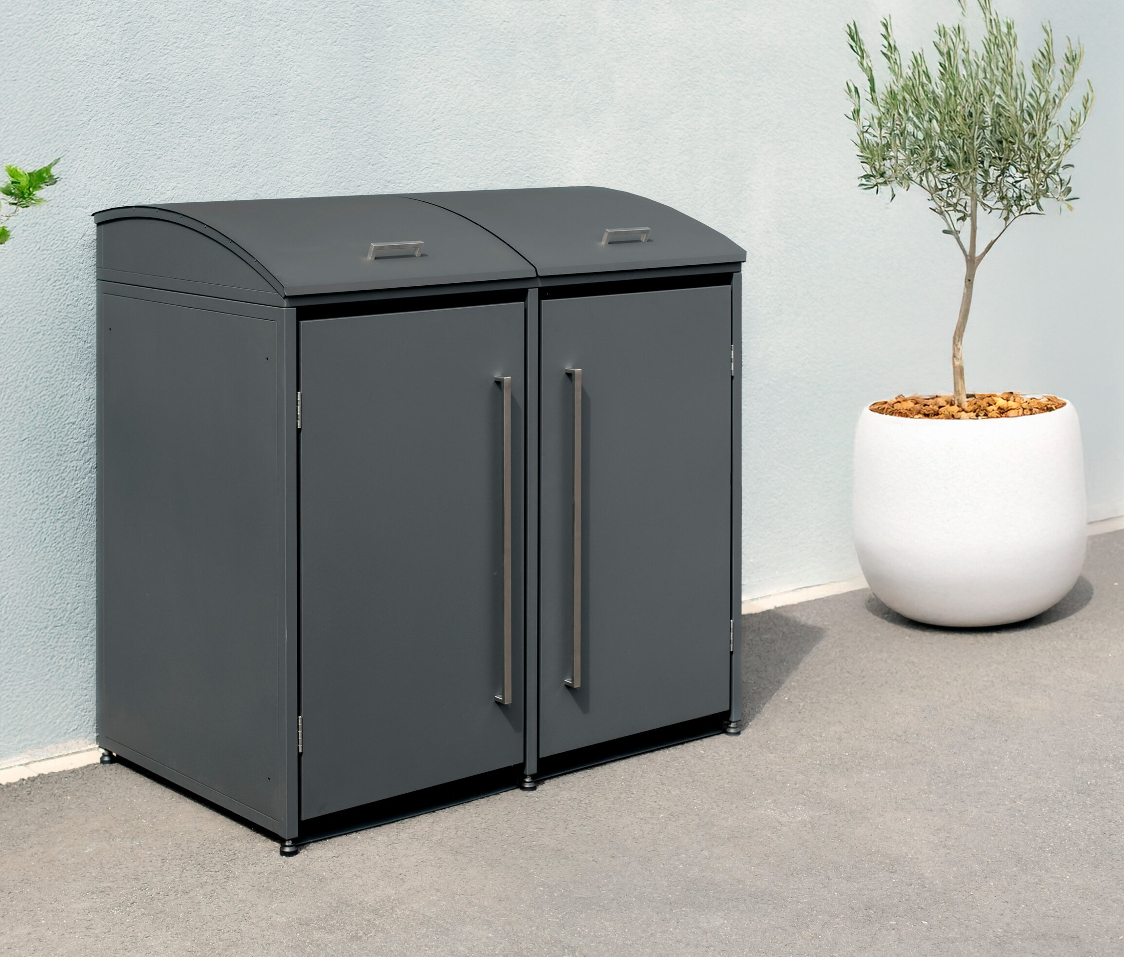 Eine graue Mülltonnenbox für zwei Mülltonnen steht draußen neben einem Baum in einem Topf.