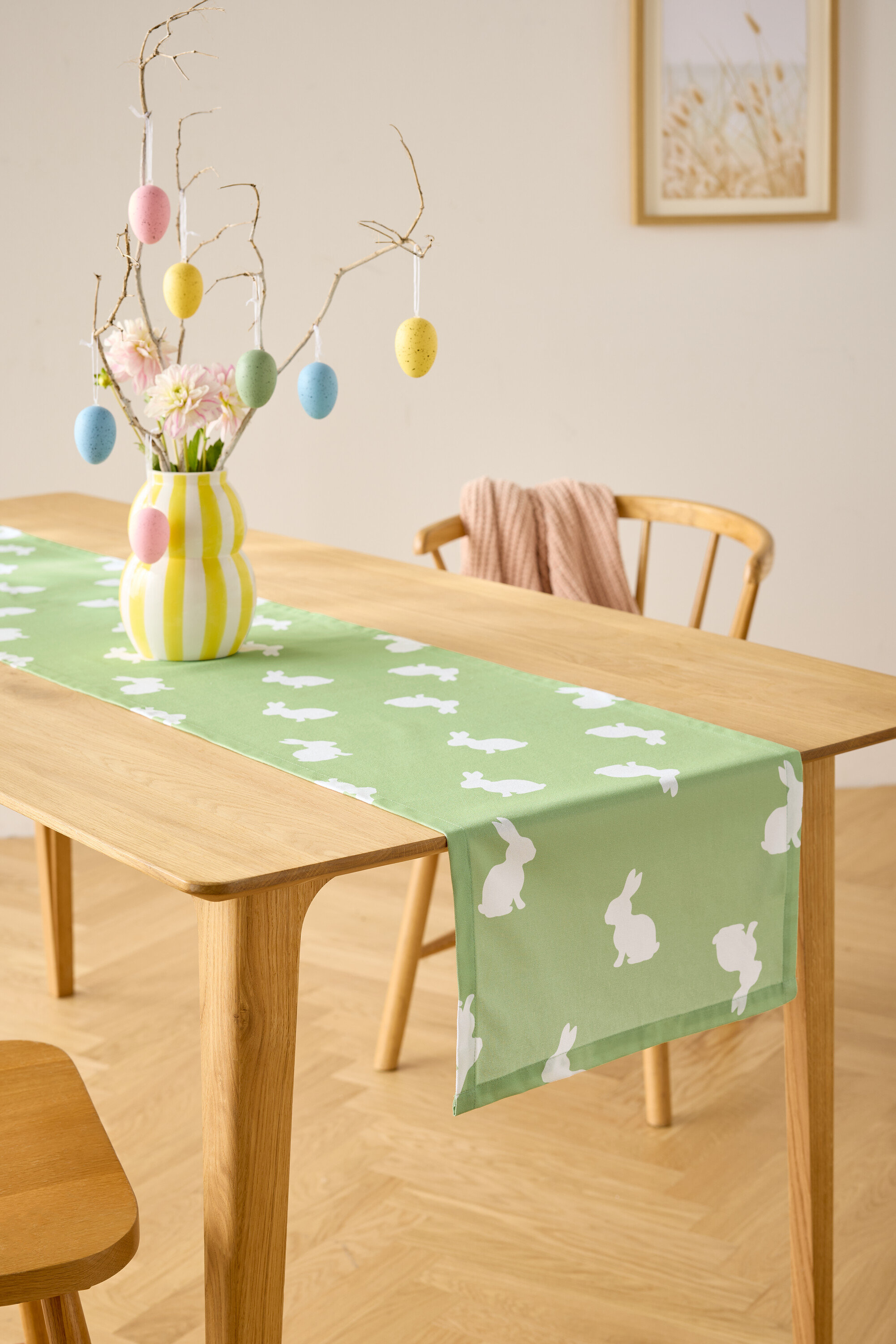 Esstisch mit hellgrünem Tischläufer mit weißen Hasenmotiven, Vase mit Blumen und Ostereier-Dekoration.
