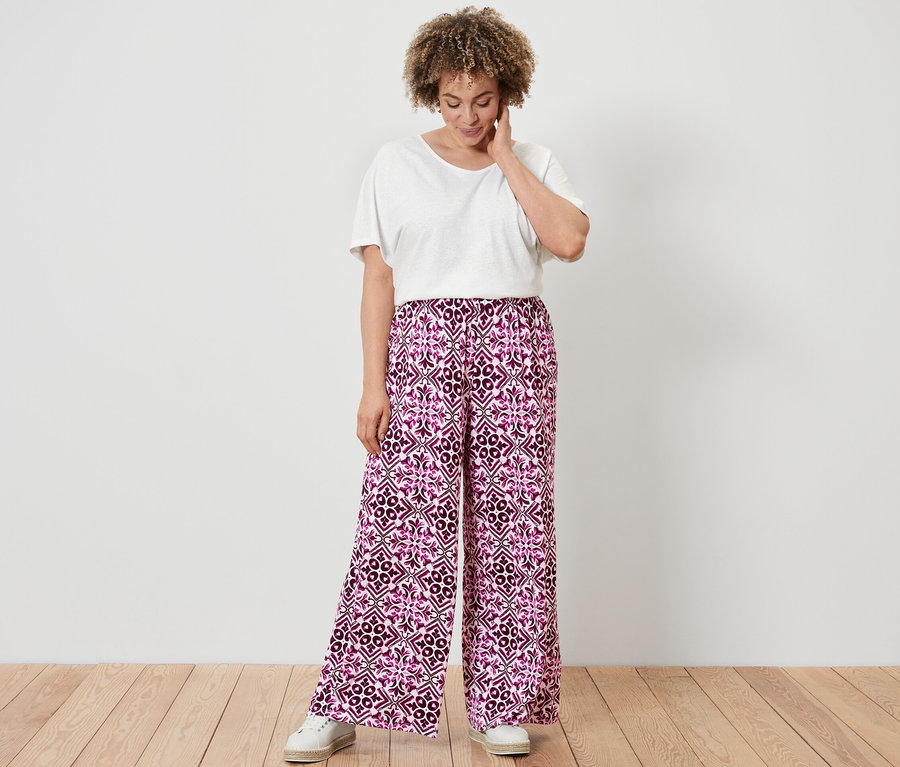 Eine Frau in einem weißen Shirt aus Leinenmix und einer bedruckten Palazzohose in Berry steht auf einem Holzboden.