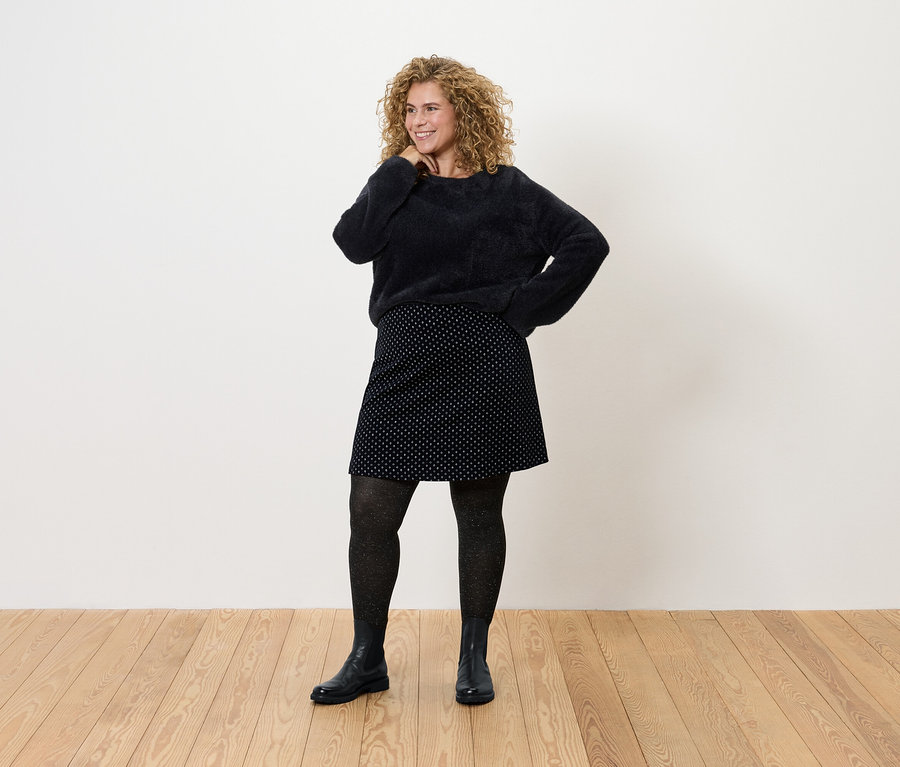 Frau mit lockigem Haar posiert in schwarzem Pullover, gepunktetem Rock, Strumpfhosen und Stiefeln.