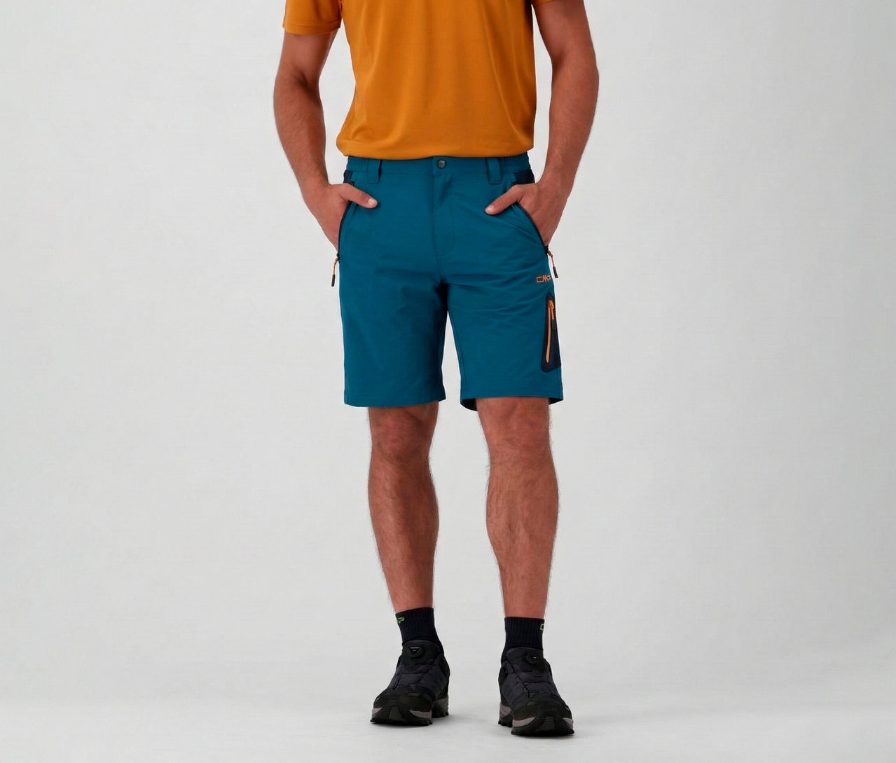 Mann mit orangefarbenem T-Shirt und CMP Herren-4-Wege-Stretch-Bermuda-Shorts in Petrol mit Händen in den Taschen.