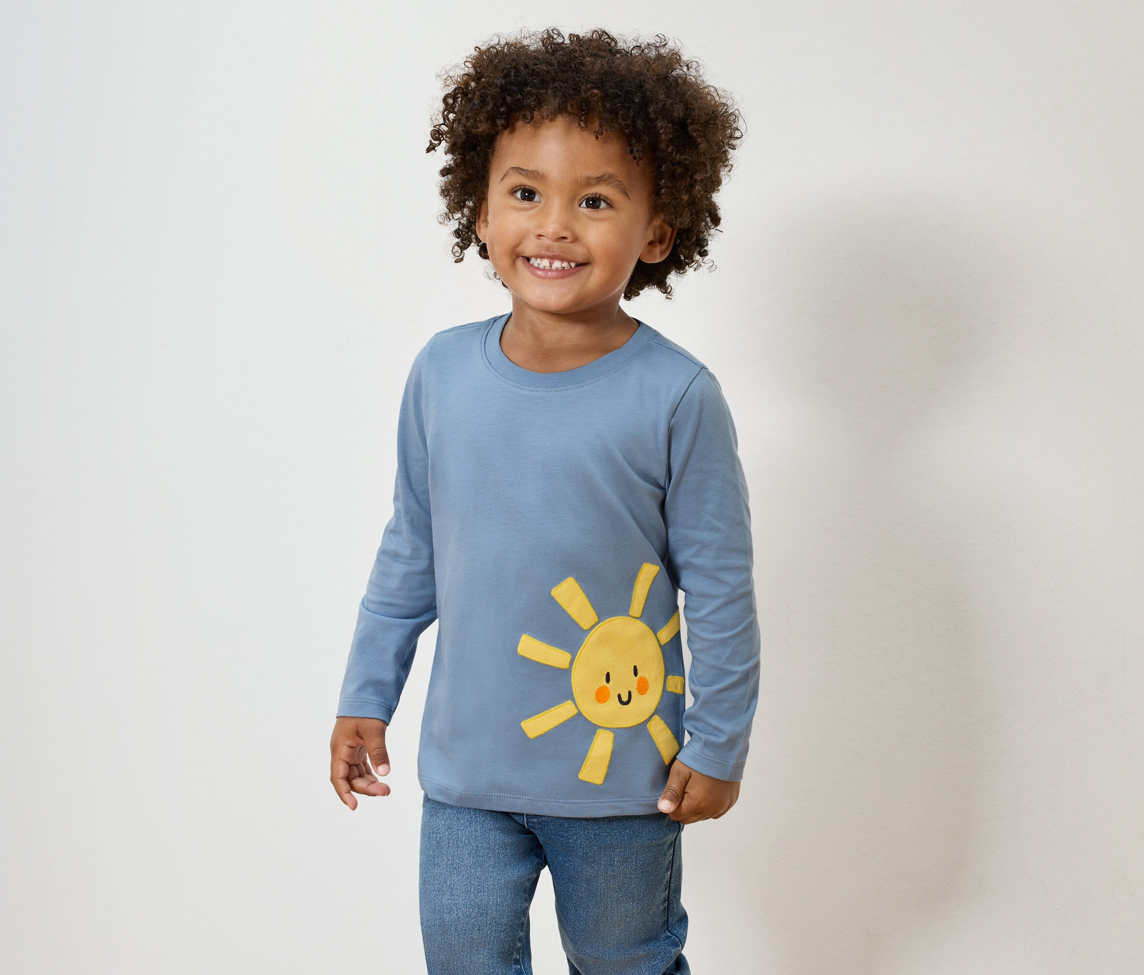Ein lächelnder Junge mit lockigem Haar trägt ein blaues Langarmshirt mit einer gelben Sonnenapplikation und Jeans.