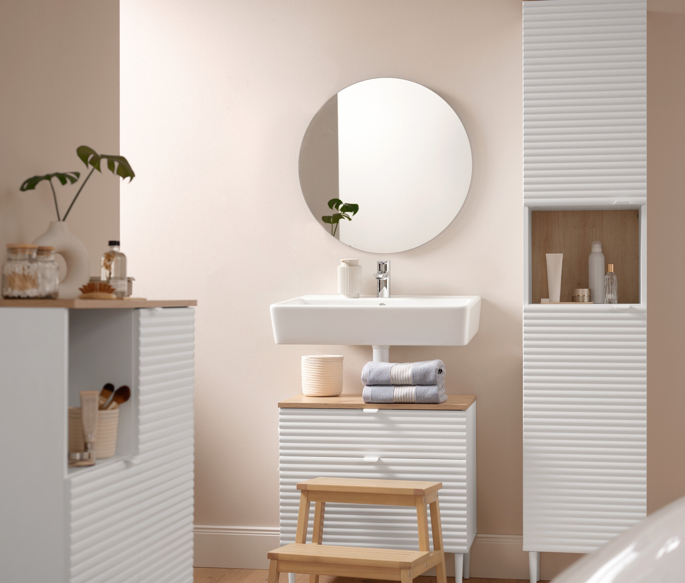 Ein Badezimmer zeigt einen Badhochschrank »SONARI« mit Türen, einen Waschbeckenunterschrank »SONARI« mit Schubladen und eine Badkommode »SONARI« mit Türen. Eine Komfort-Badematte (60 x 100 cm) liegt vor einem Tritthocker aus Massivholz. Auf dem Waschbeckenunterschrank liegen zwei blaue Premium-Handtücher. Ein runder Spiegel »Ameca« hängt über dem Waschbecken.