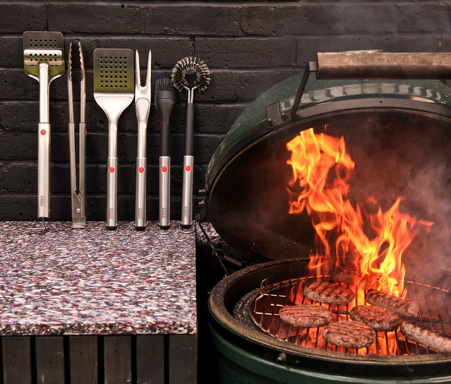 Grillwerkzeuge hängen neben einem grünen Keramikgrill, in dem Burger über offenem Feuer braten.
