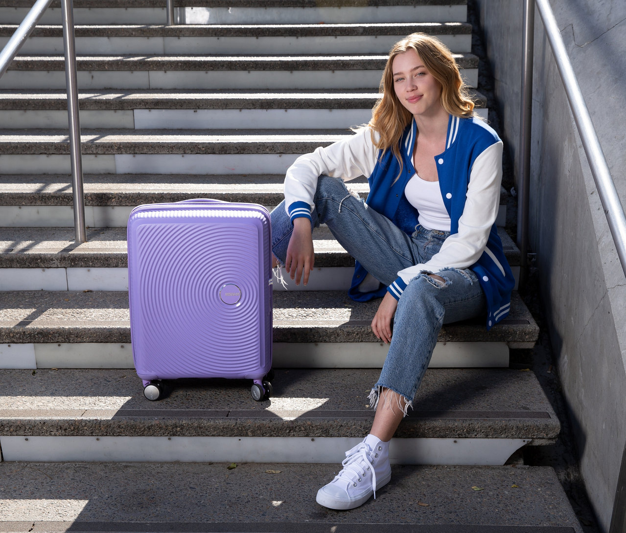 Frau sitzt auf Treppe neben American Tourister Hartschalen-Koffer »Soundbox« Spinner, klein, lavender.