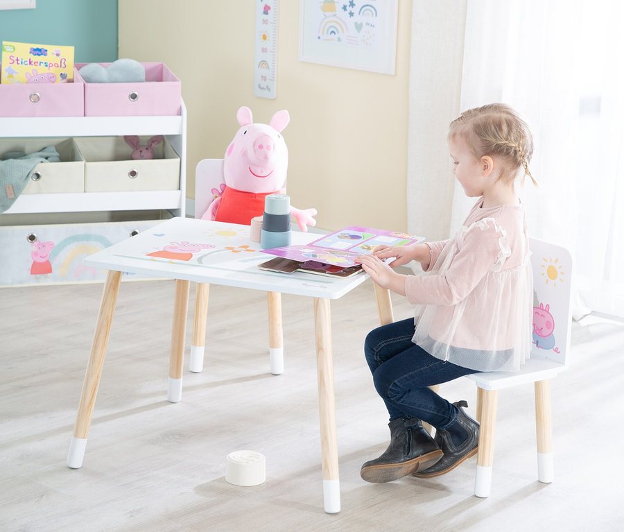 Mädchen sitzt an einem Tisch mit Peppa Pig-Motiven und betrachtet ein Buch. Ein Peppa Pig Plüschtier sitzt auf einem Stuhl neben dem Tisch.