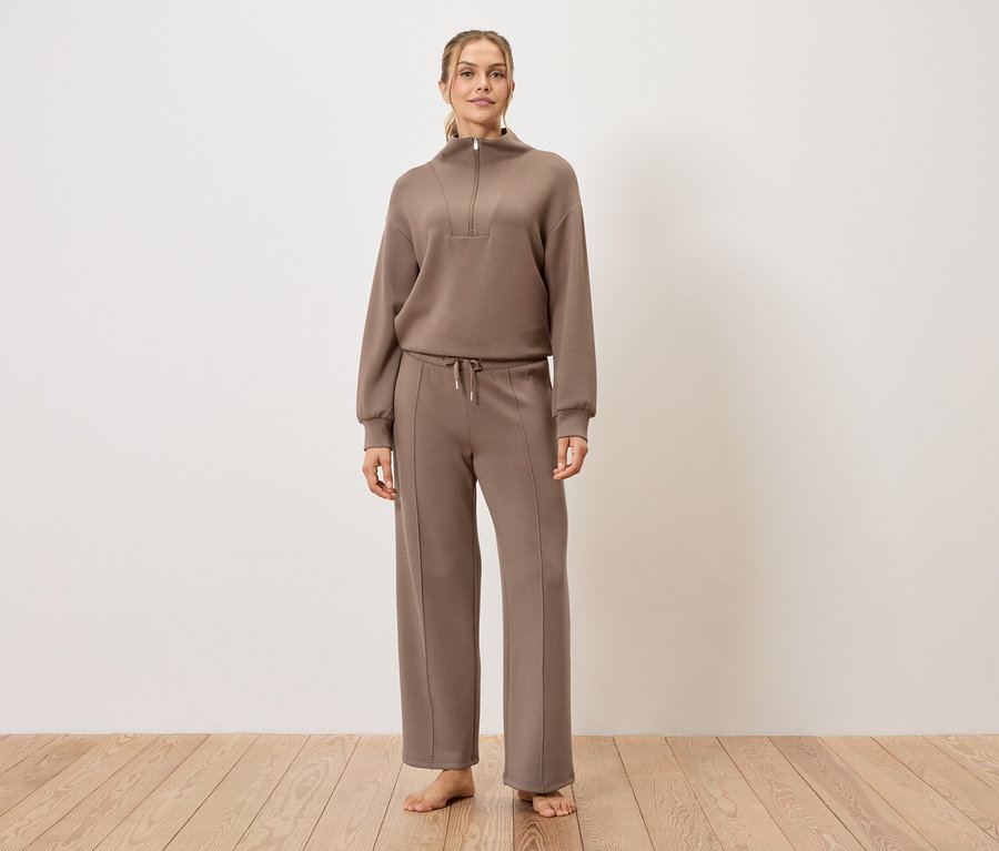 Frau steht barfuß auf Holzboden und trägt ein braunes Trainingsanzug-Set.